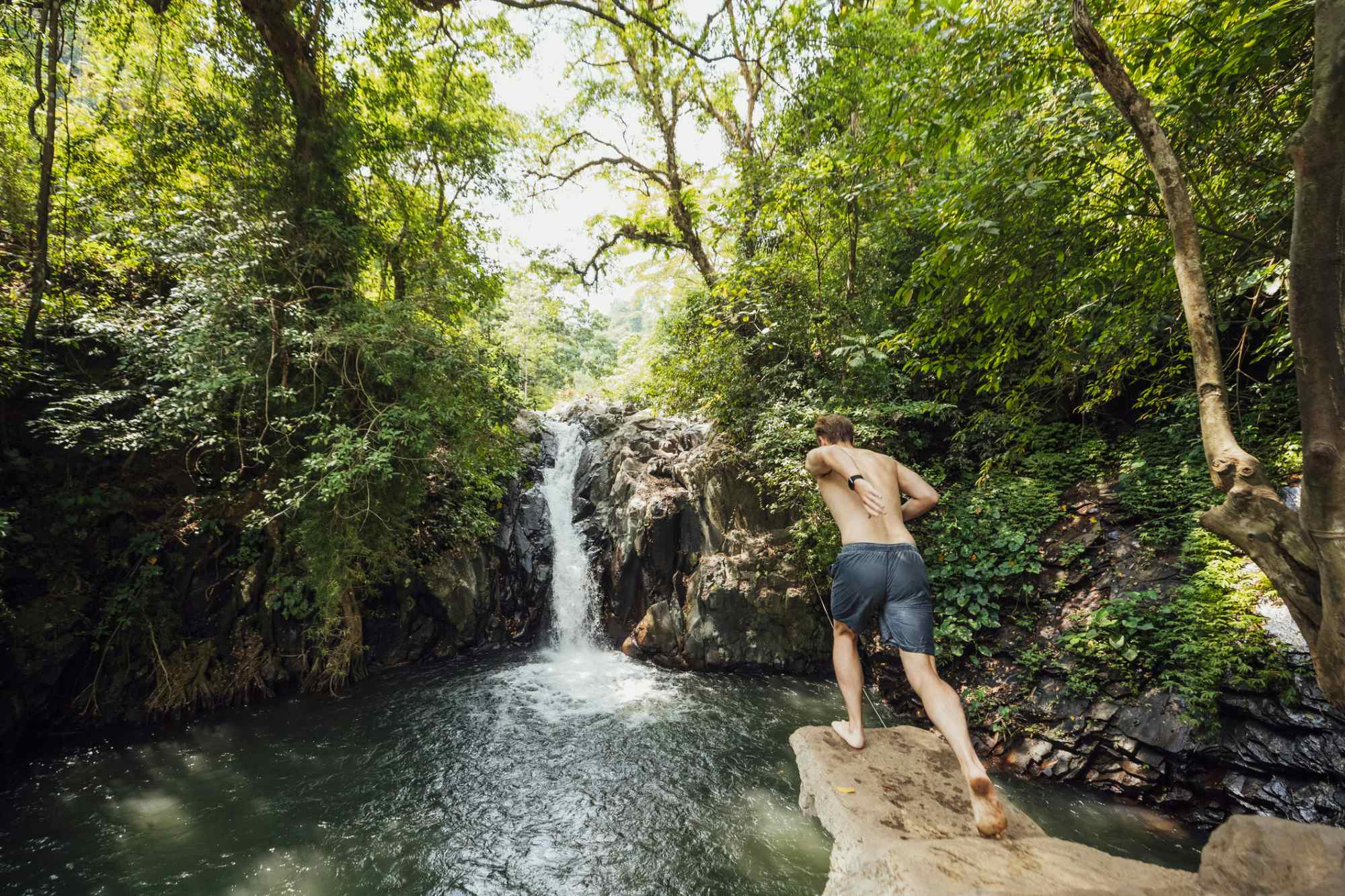 A rear view action shot of a young man taking a running leap off a cliff next to a waterfall in Bali.
