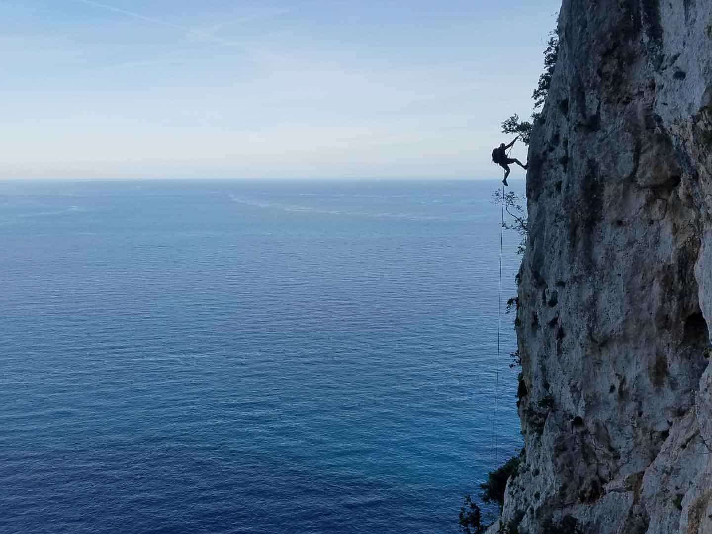 Abseiler on a cliff silhouetted against the blue sea on the Selvaggio Blu trail in Sardinia, Italy.