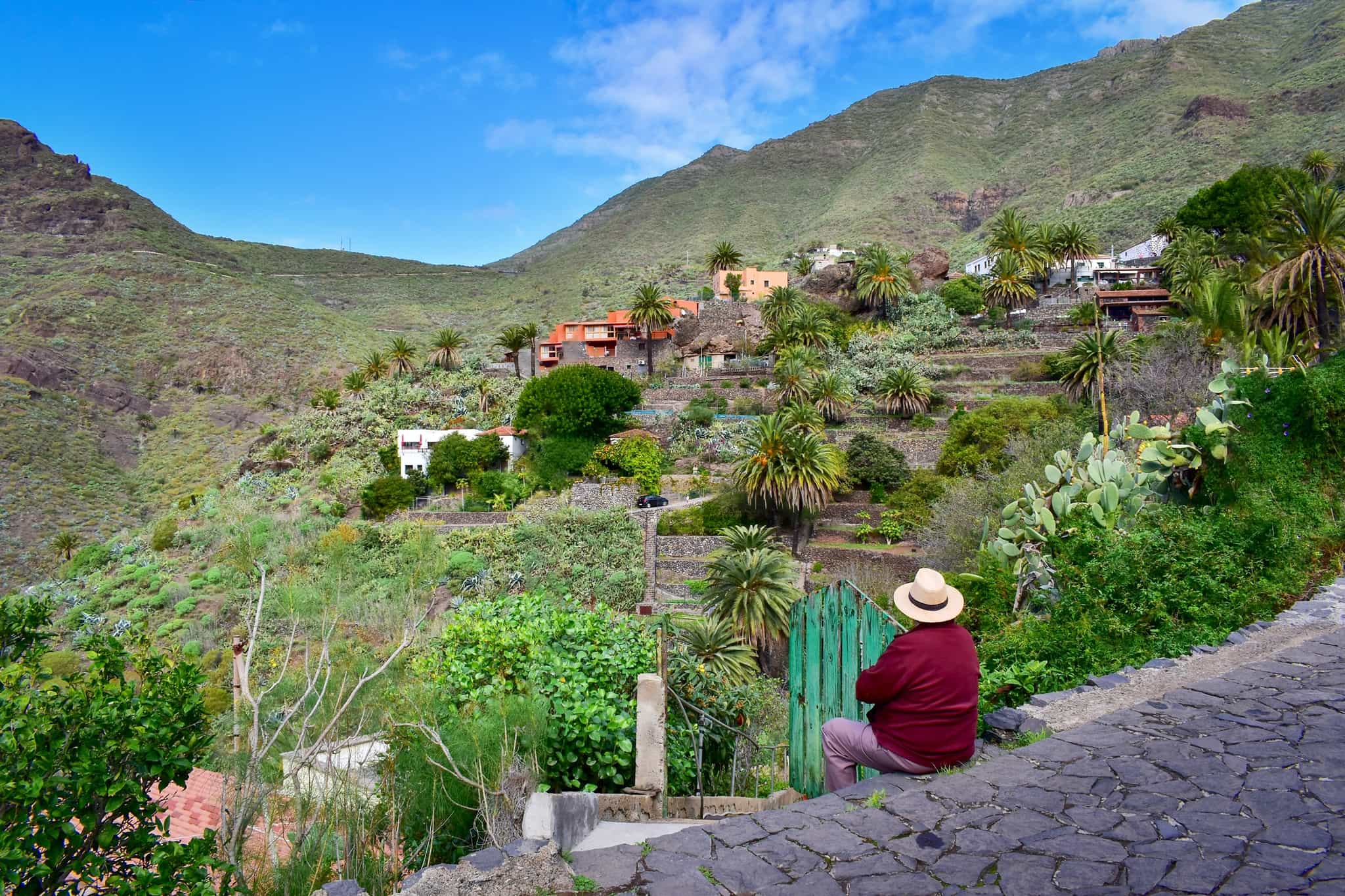 Man wearing a hat and red jumper sits on a wall and looks up at a small village in the hills of Tenerife.