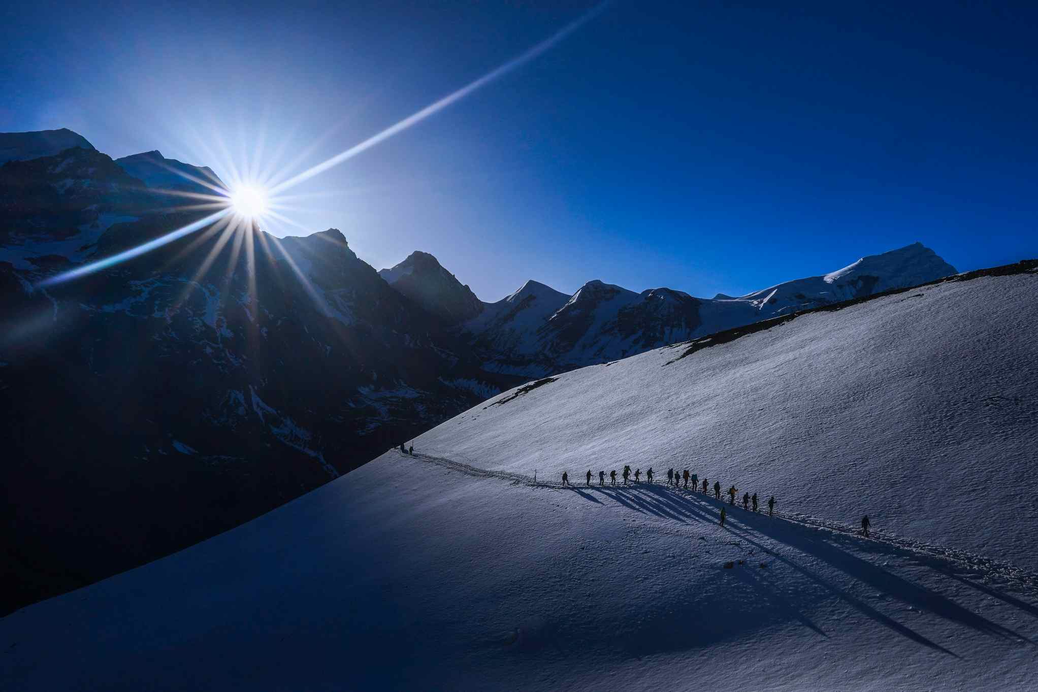 A string of trekkers on the Thorong La Pass, with the sun rising over the mountains in the background, Nepal. 