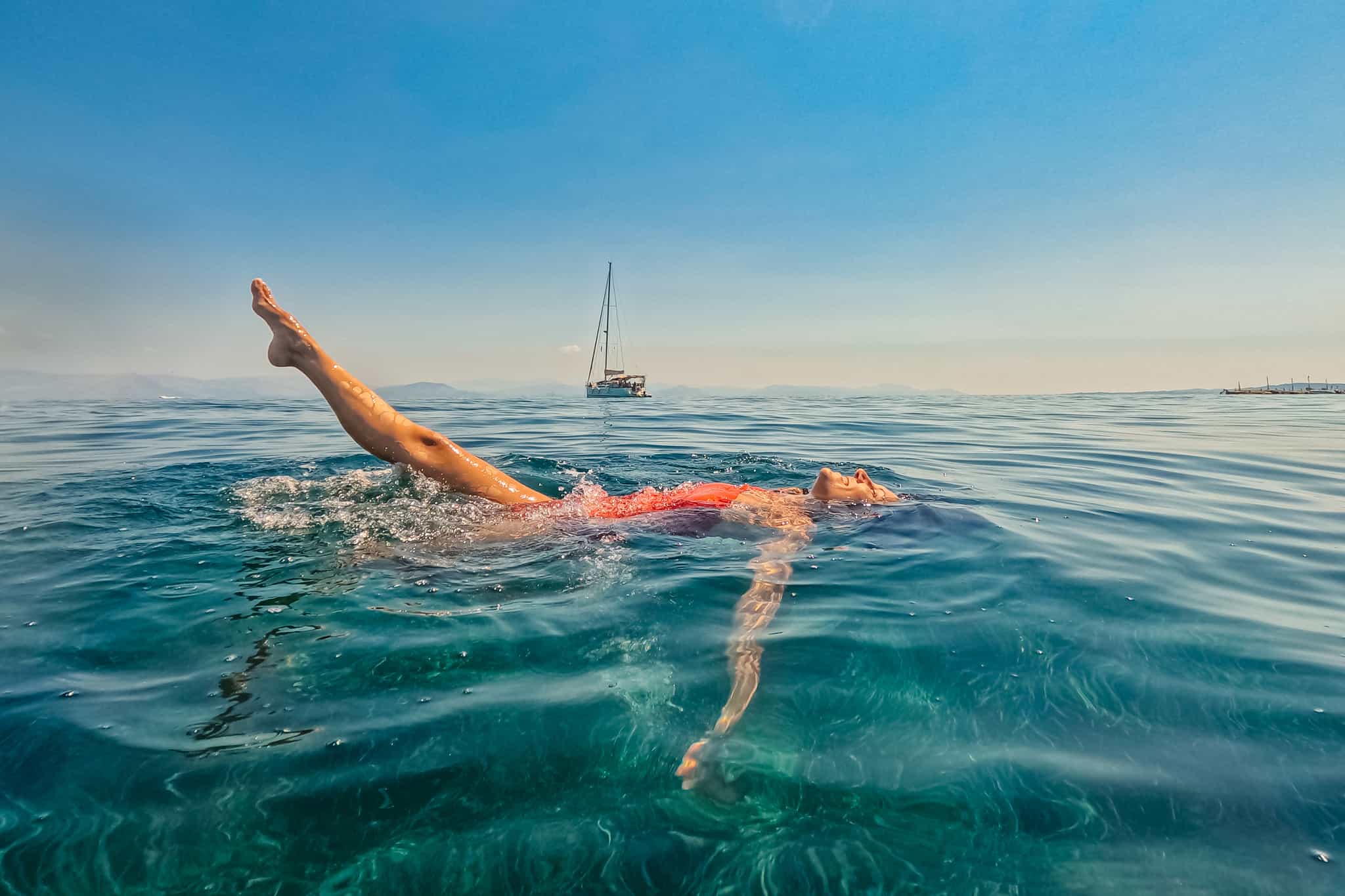 Woman swimming with a sailboat in the background, Greece.
