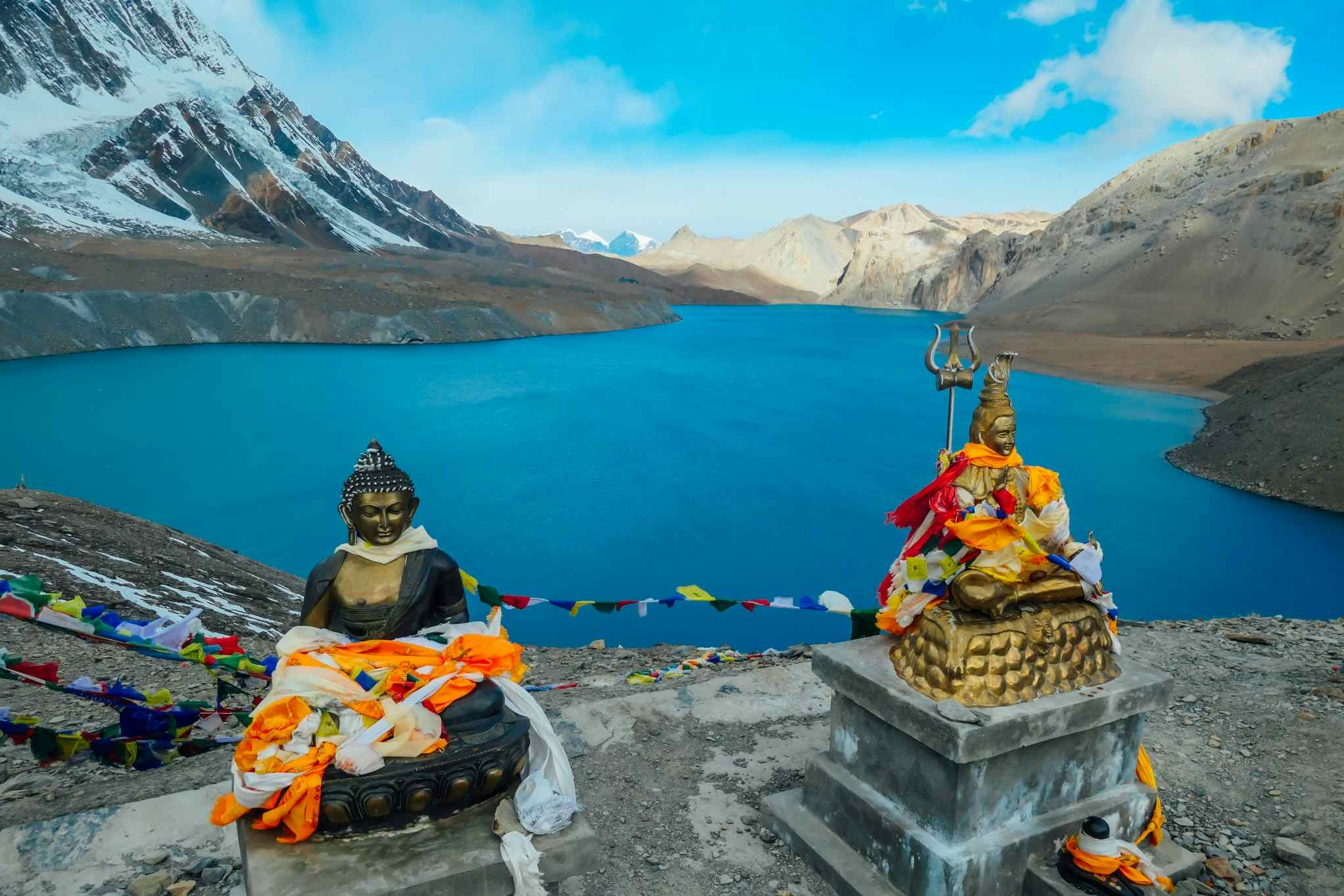 Two Buddha statues at Tilicho Lake, covered with prayer flags, backed by a blue lake and mountains on the Annapurna Circuit Trek, Nepal.