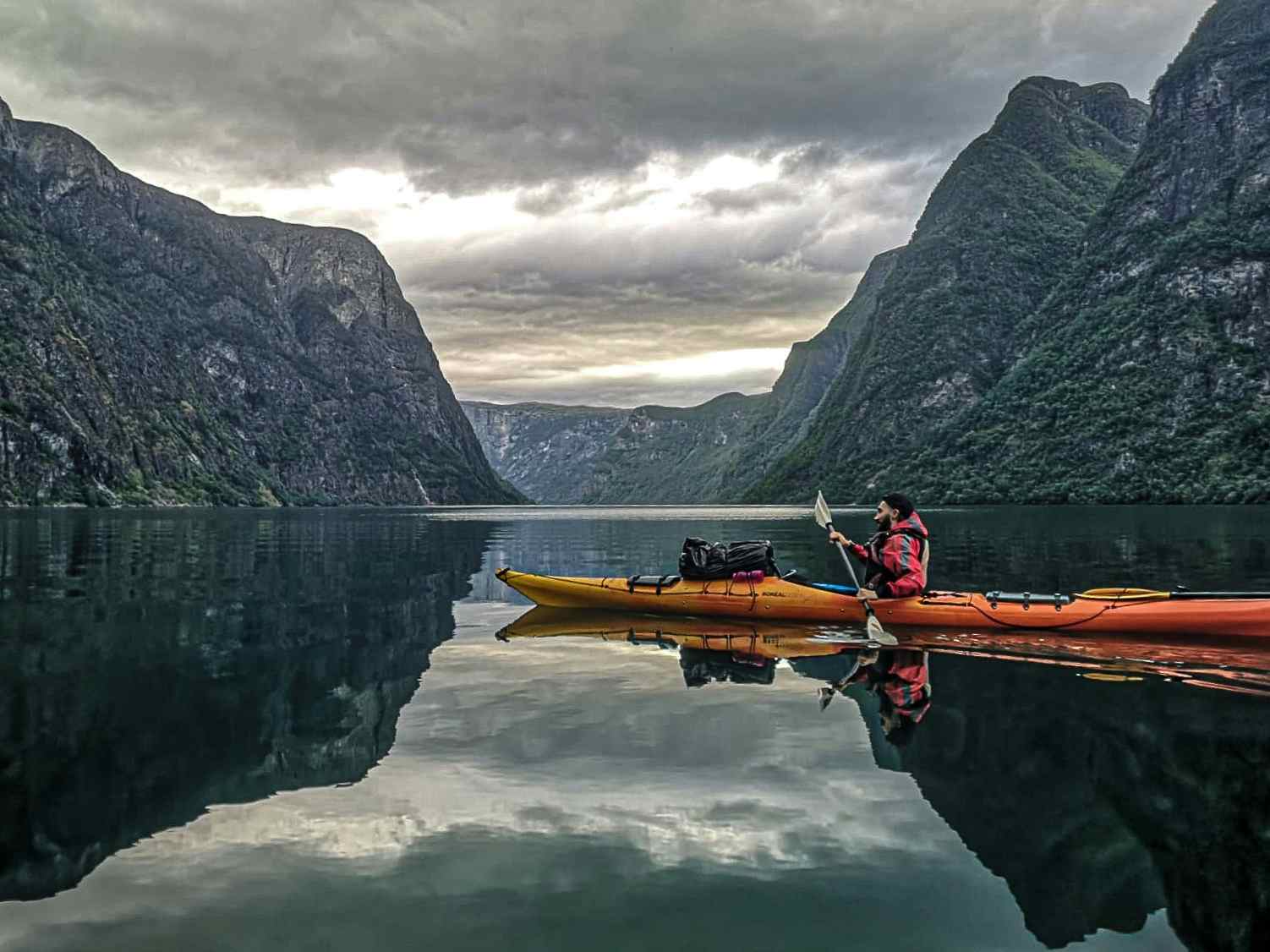 A kayak paddles along the serene waters of the Naeroyfjord in the Norwegian Fjords on a cloudy day. 