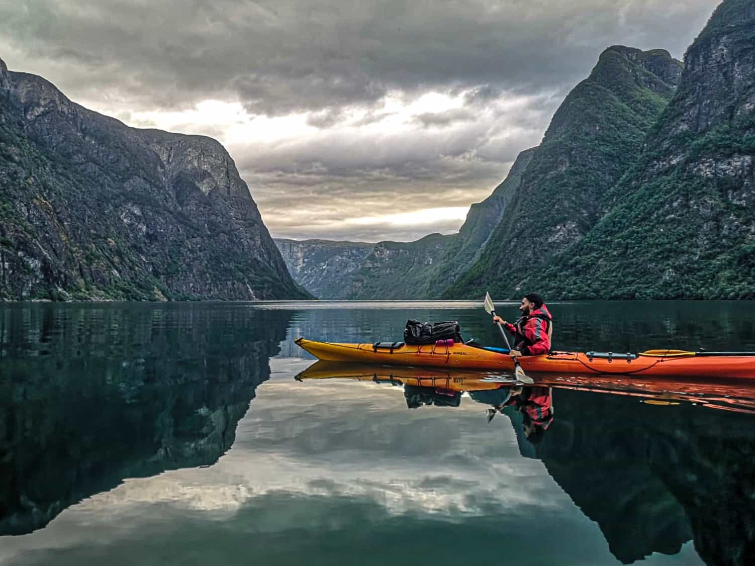 A kayak paddles along the serene waters of the Naeroyfjord in the Norwegian Fjords on a cloudy day.