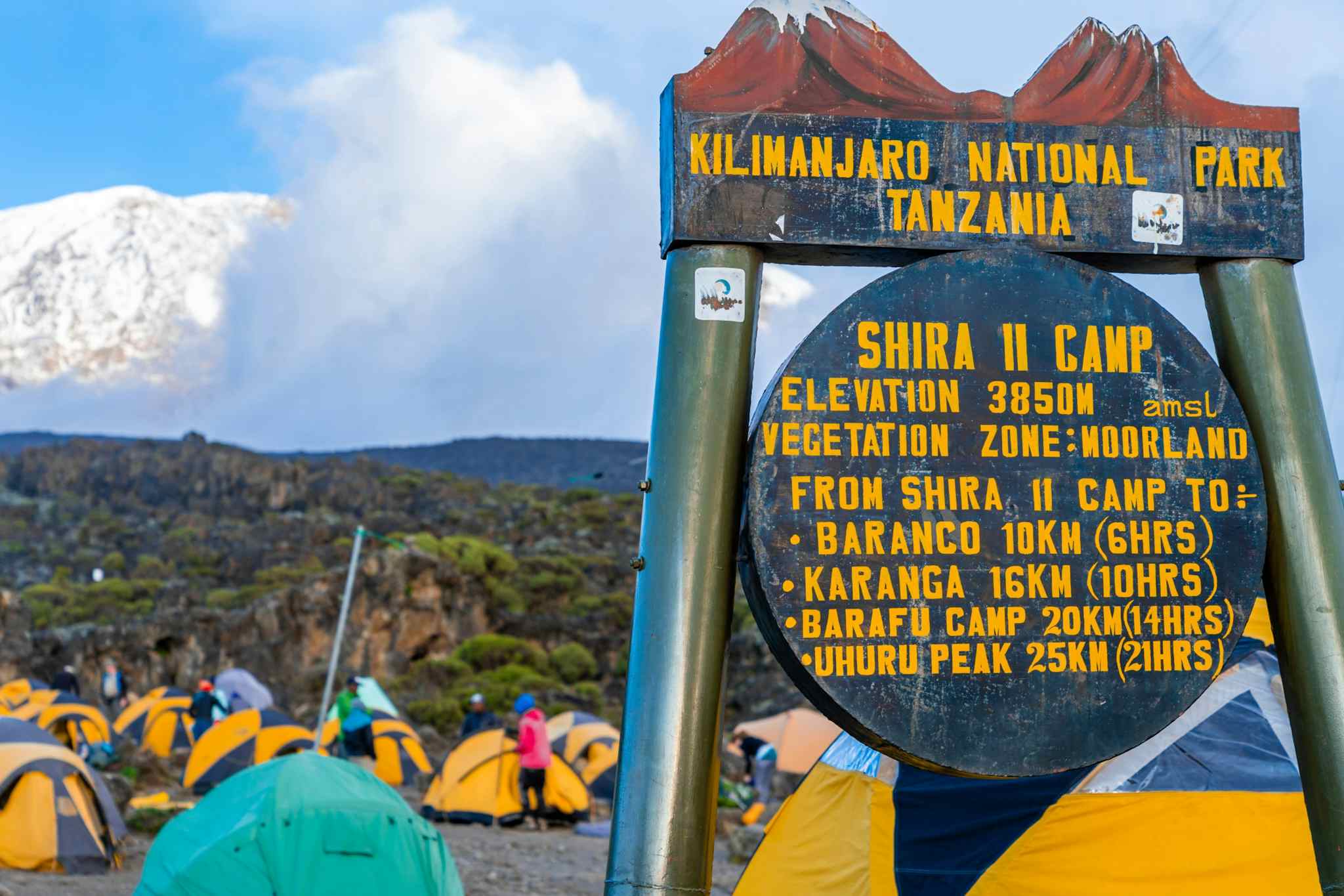 The signpost for Shira Camp II, with tents and Mount Kilimanjaro's summit in the background.