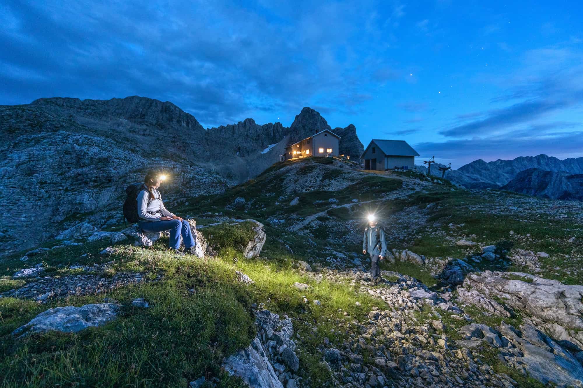 Two hikers exploring by headtorch at dusk outside the Kredarica Mountain Hut in the Julian Alps