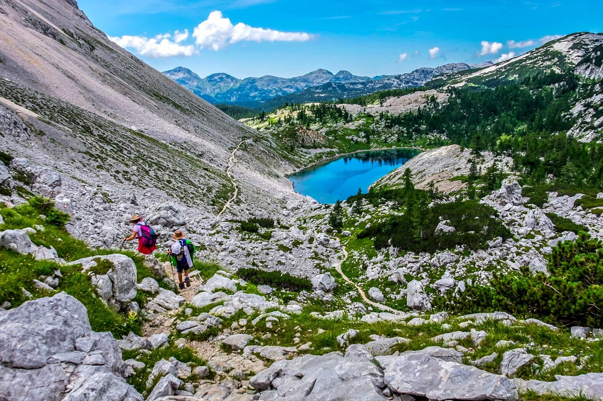Two hikers making their way down a mountain trail past a lake