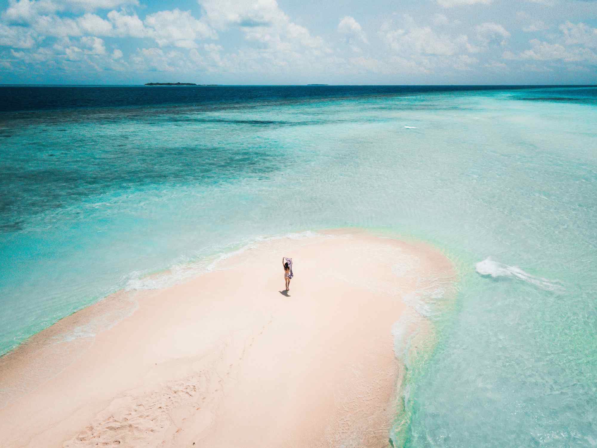 An aerial photograph of a person stood on a white sandbar surrounding by clear, turquoise, water.