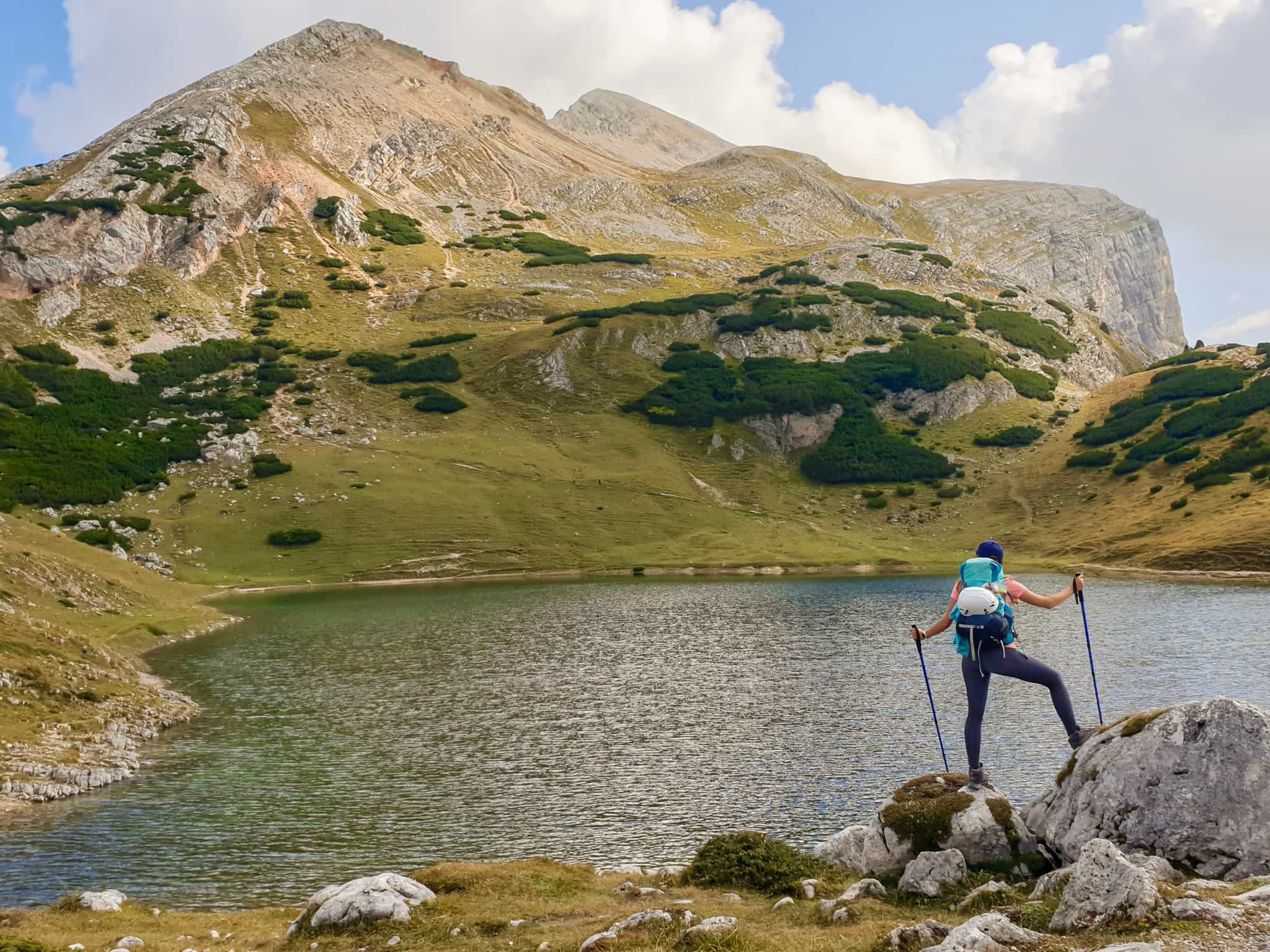 A woman with a big hiking backpack standing on a big boulder at the shore of Le de Limo in Italian Dolomites.