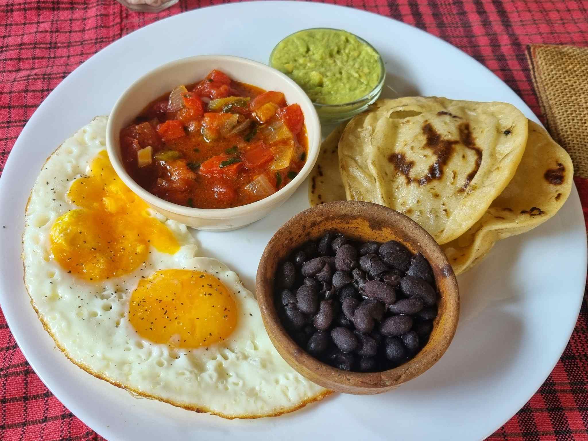 Traditional Guatemalan breakfast of eggs, black beans, & fried plantain with tortillas wrapped in basket on handwoven Maya textile.