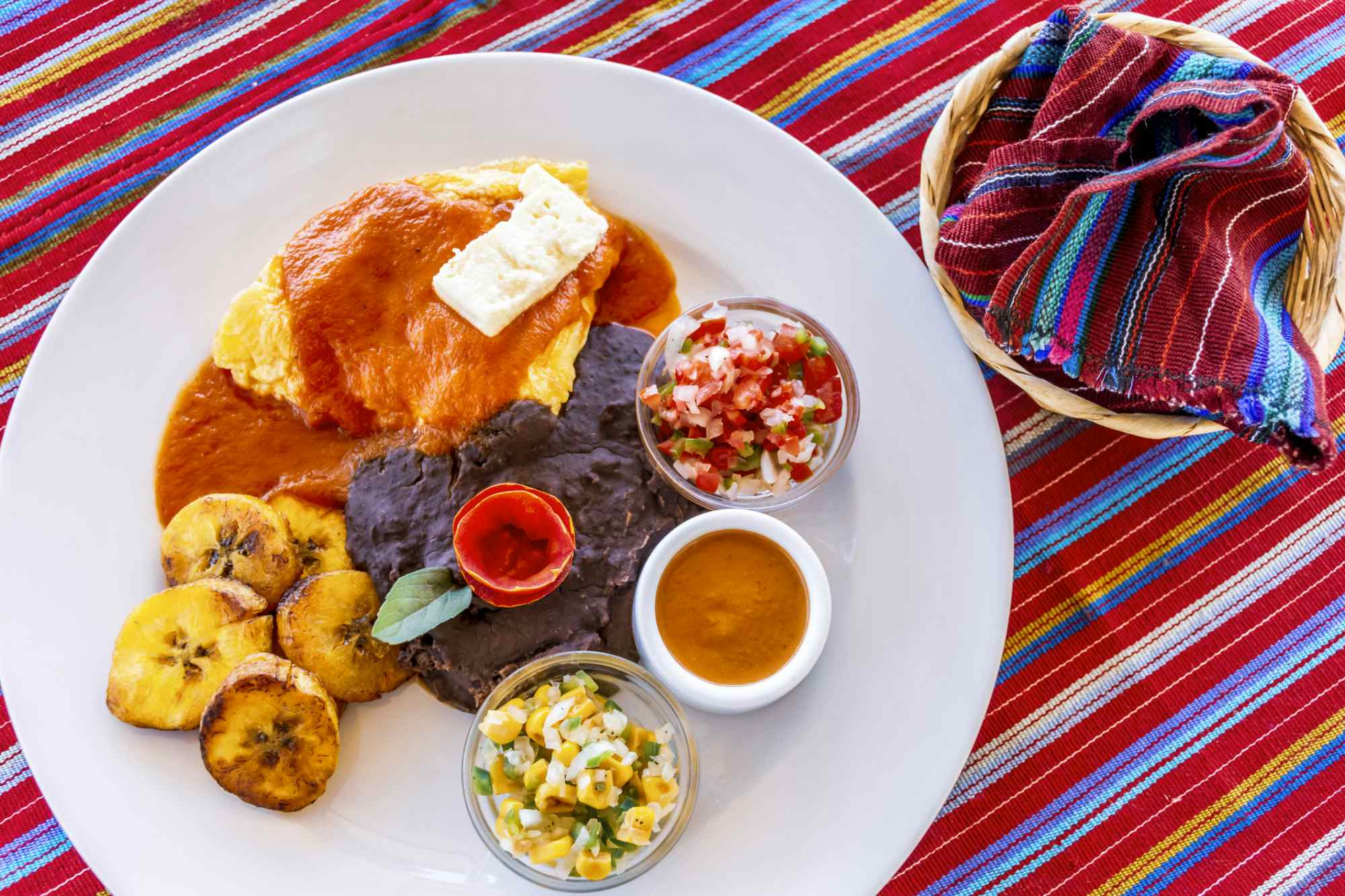 Traditional Guatemalan breakfast of eggs, black beans, cheese & fried plantain with tortillas wrapped in basket on handwoven Maya textile.