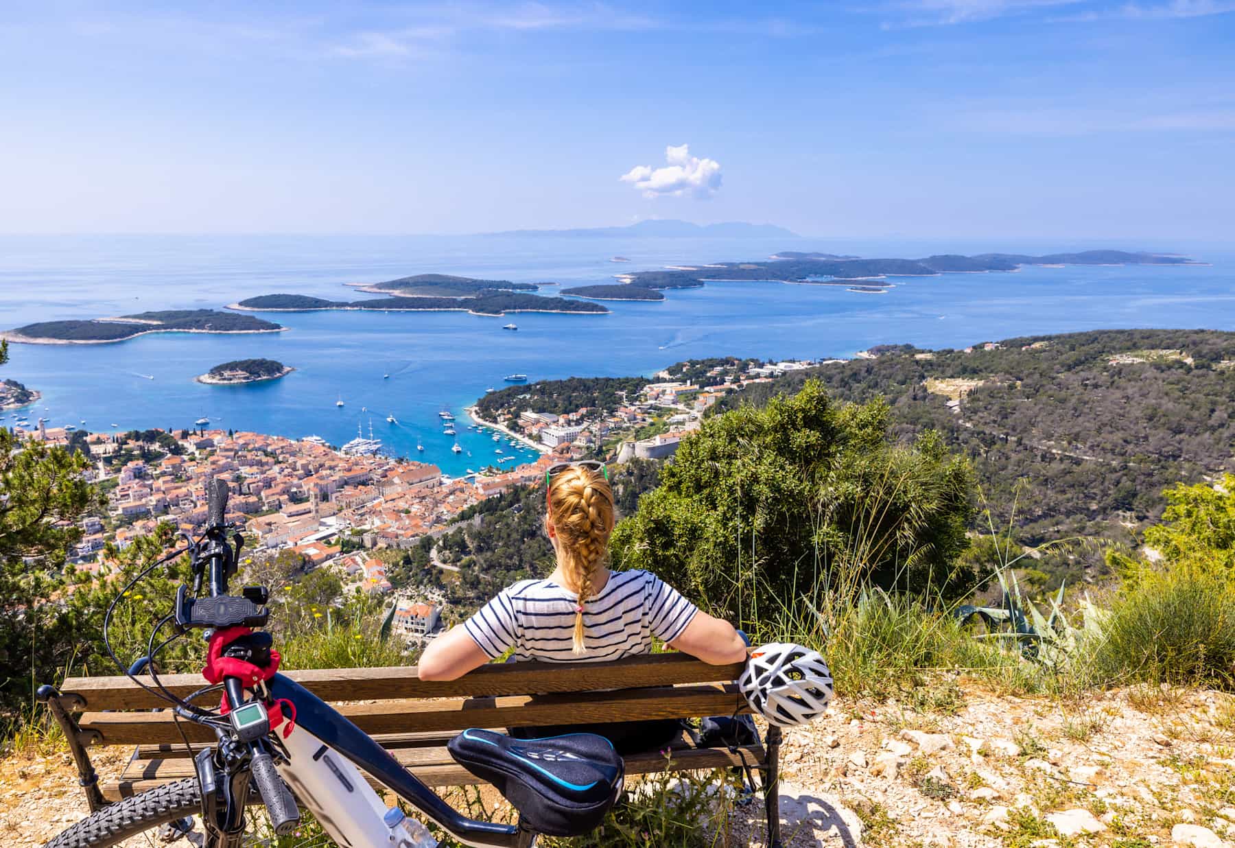 Female biker sitting on wooden bench while looking at beautiful coastal town from mountain top on sunny day, Croatia