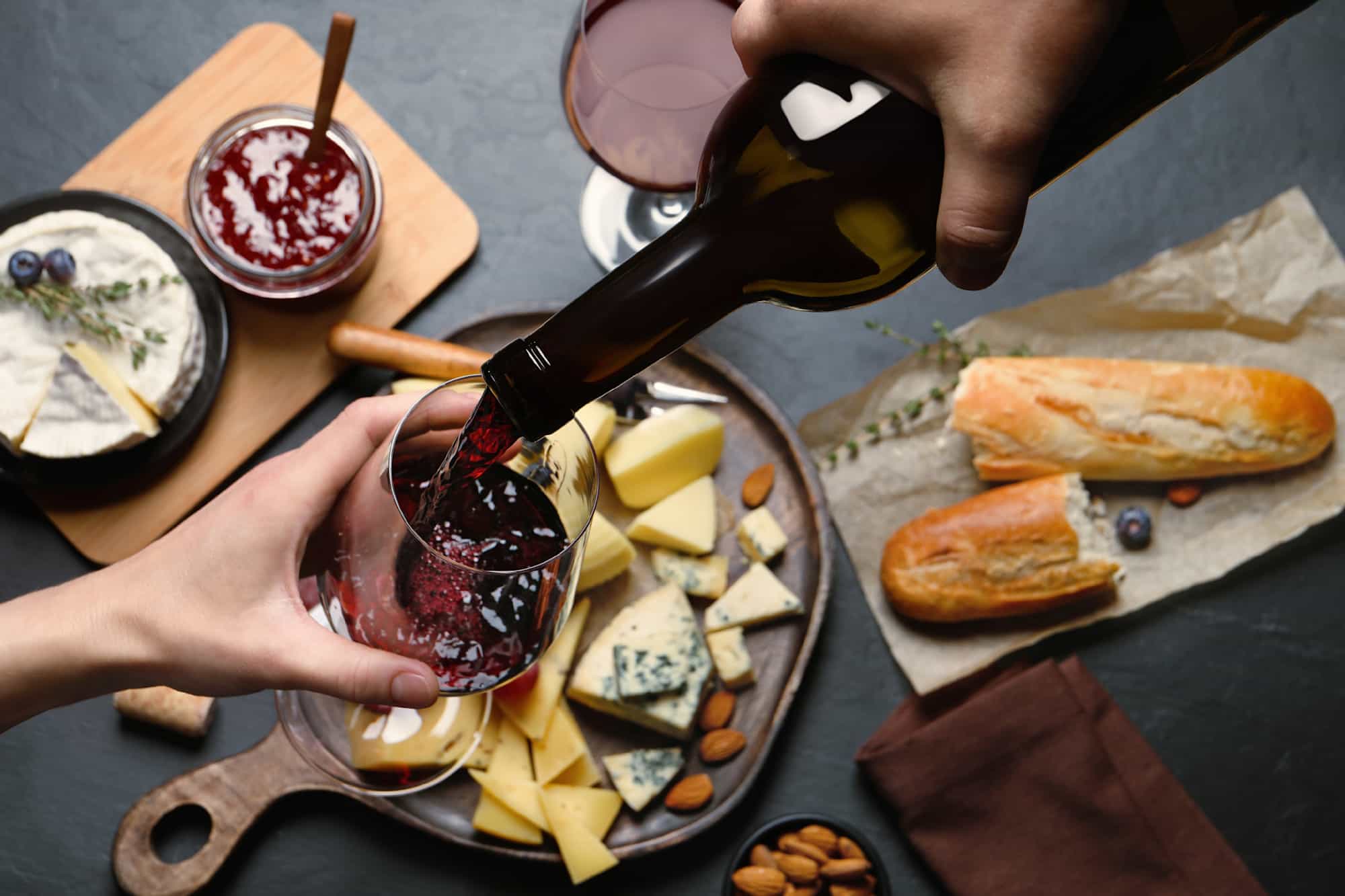 Pouring red wine from bottle into glass over black table with snacks, top view
