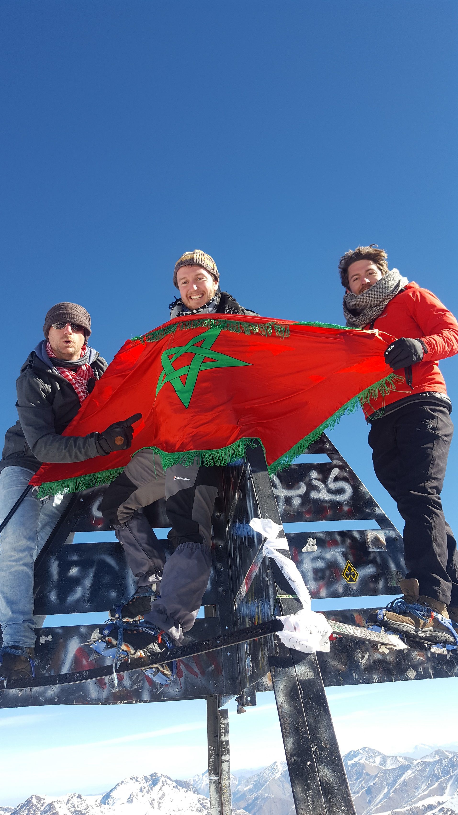 Three climbers hold a Morocco flag up on the summit of Mount Toubkal.