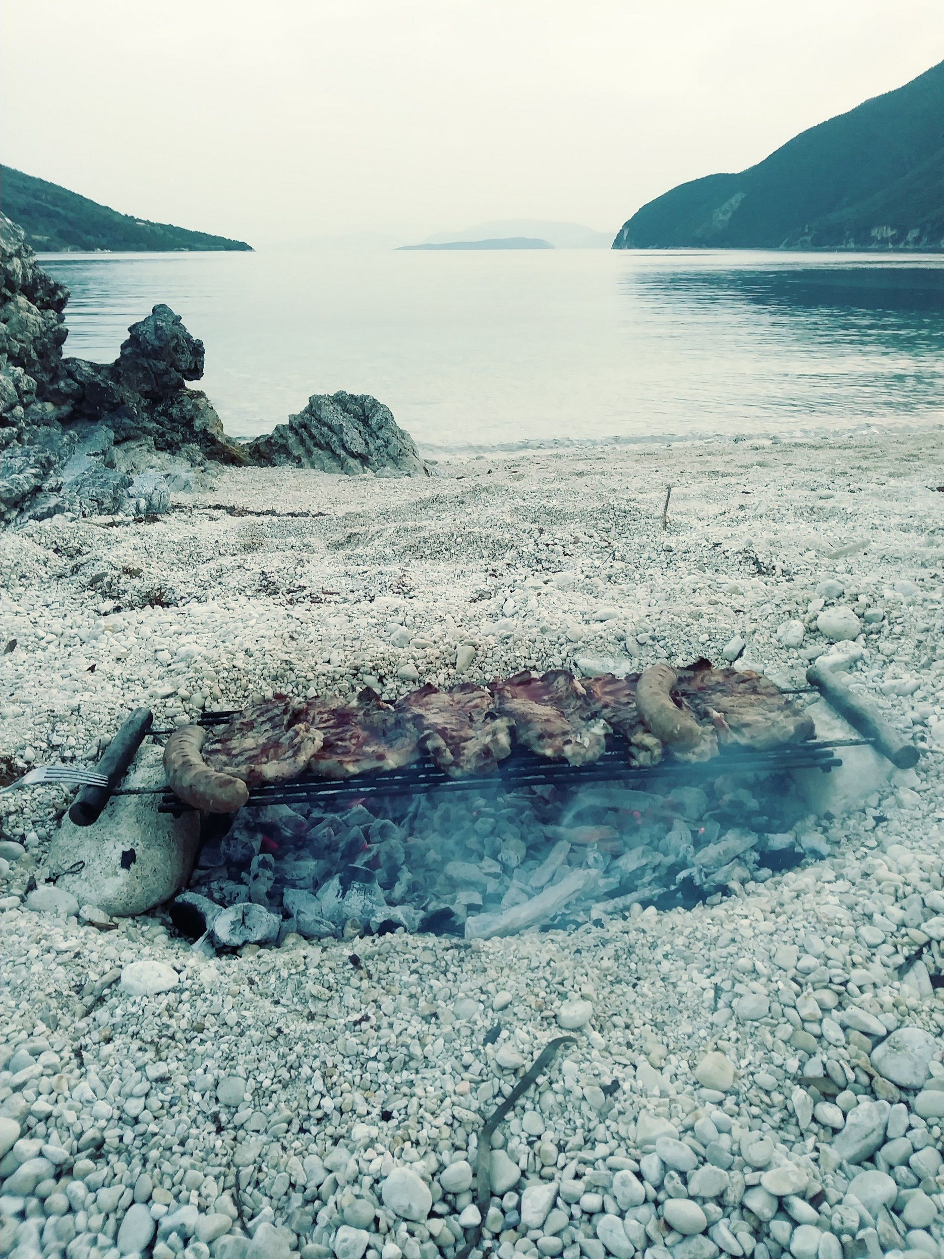 Meat and sausages being grilled over charcoal on a beach. 