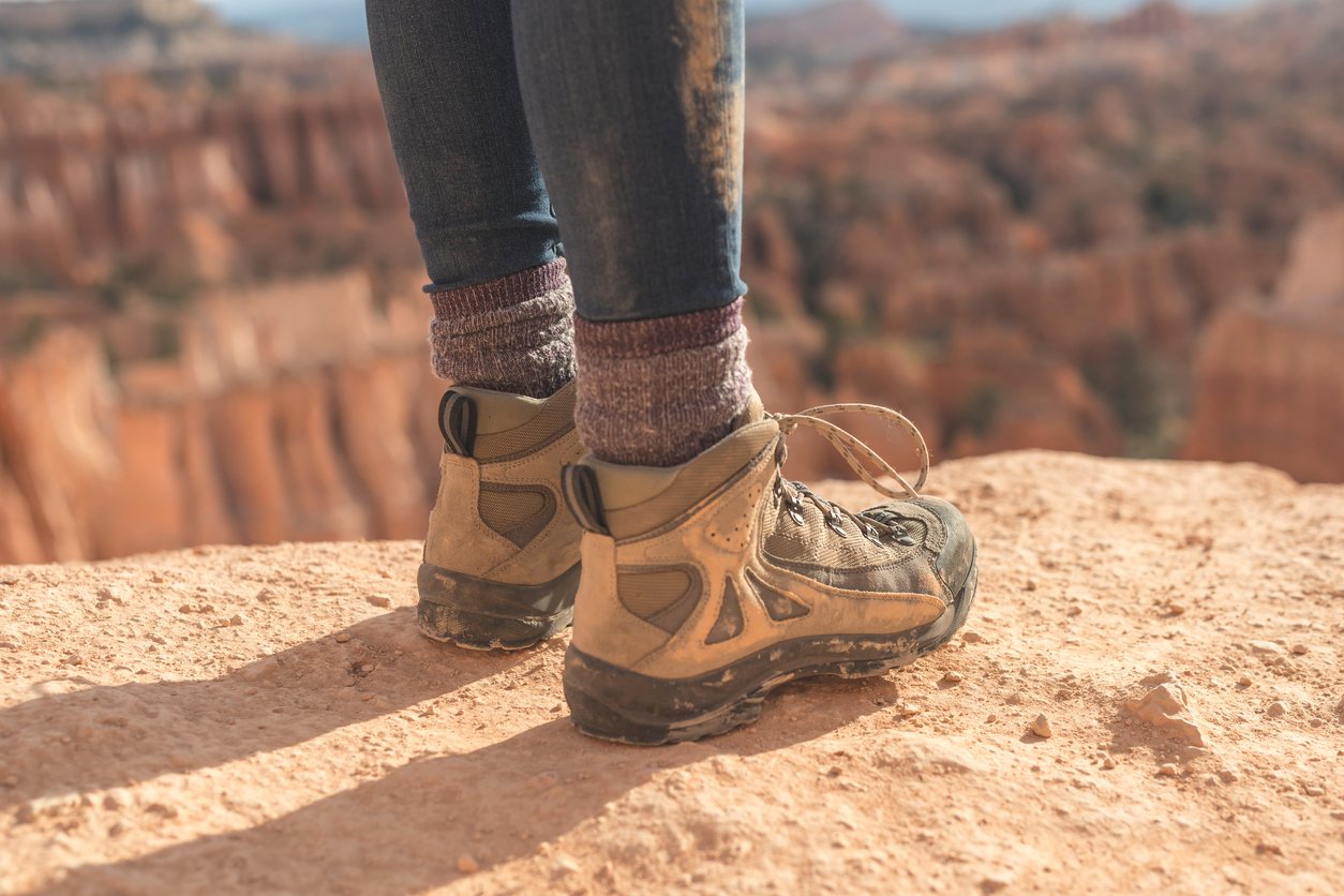 A woman stands on the edge of a cliff, with the image showing a close up of her dirty hiking boots.