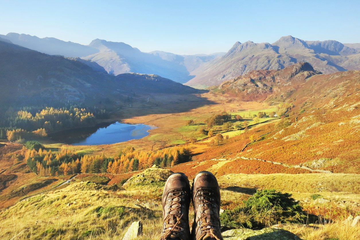 A pair of hiking boots sit in front of a beautiful view of a lake, flanked by rolling mountains.