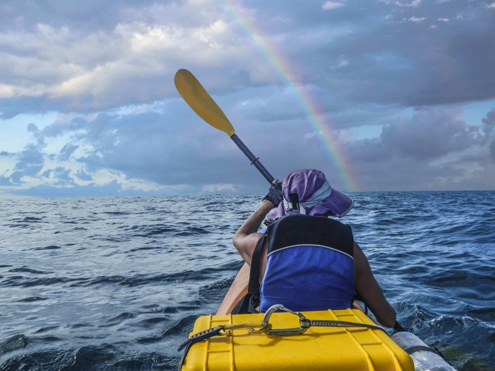 A kayaker in the ocean, with a rainbow in front of them. 