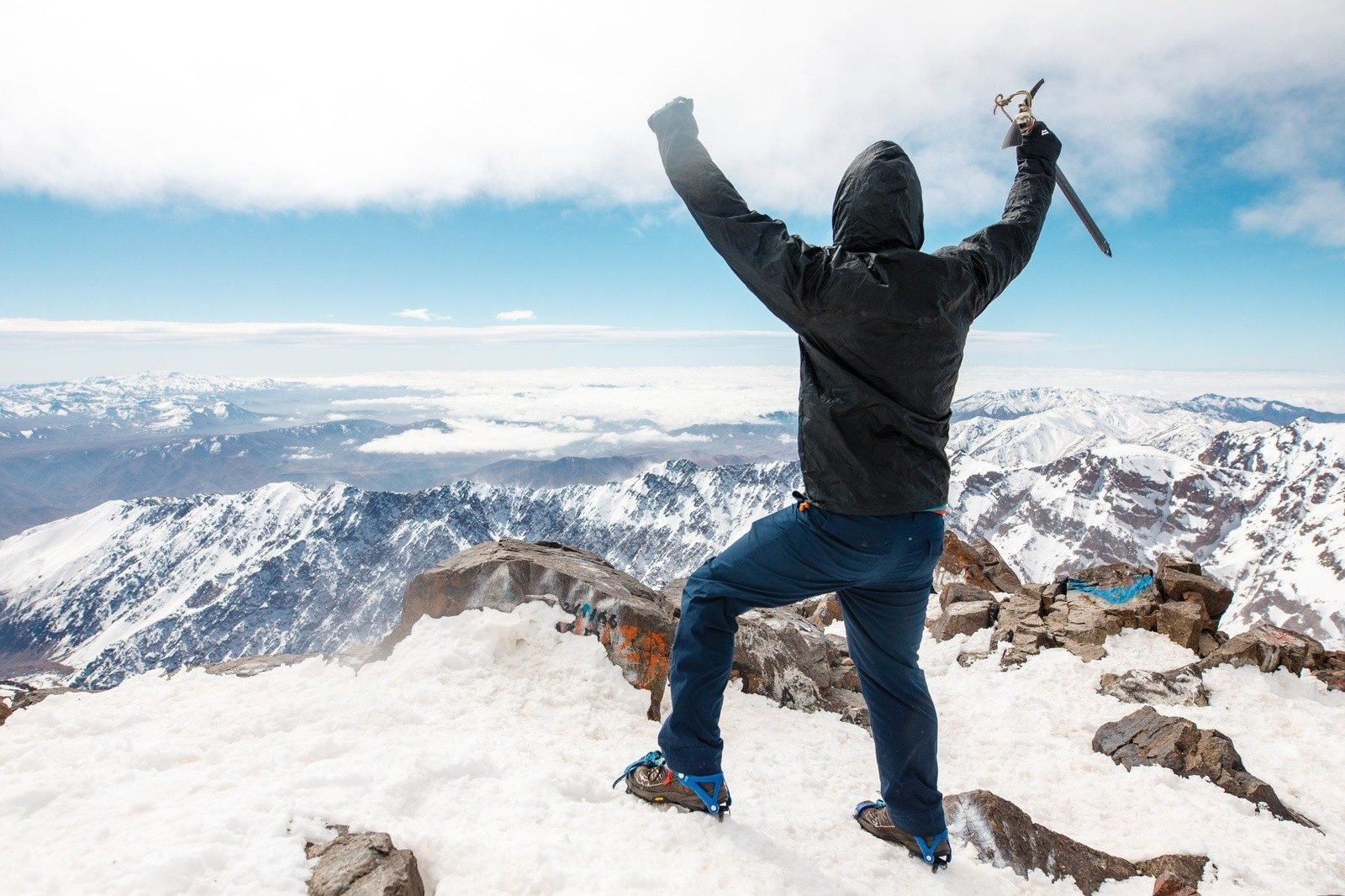 Back view of a hiker, wearing crampons, celebrating at the top of a snowy summit