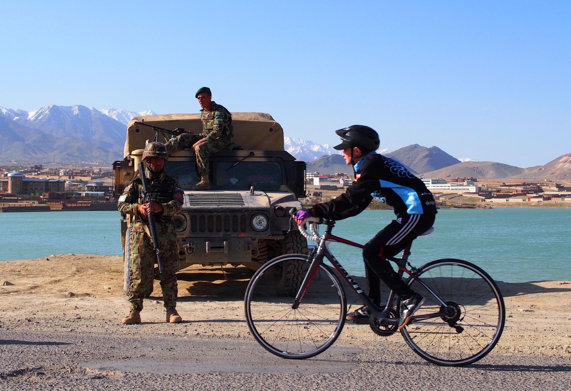 A women cycling past a military presence, in front of the mountains of Afghanistan.