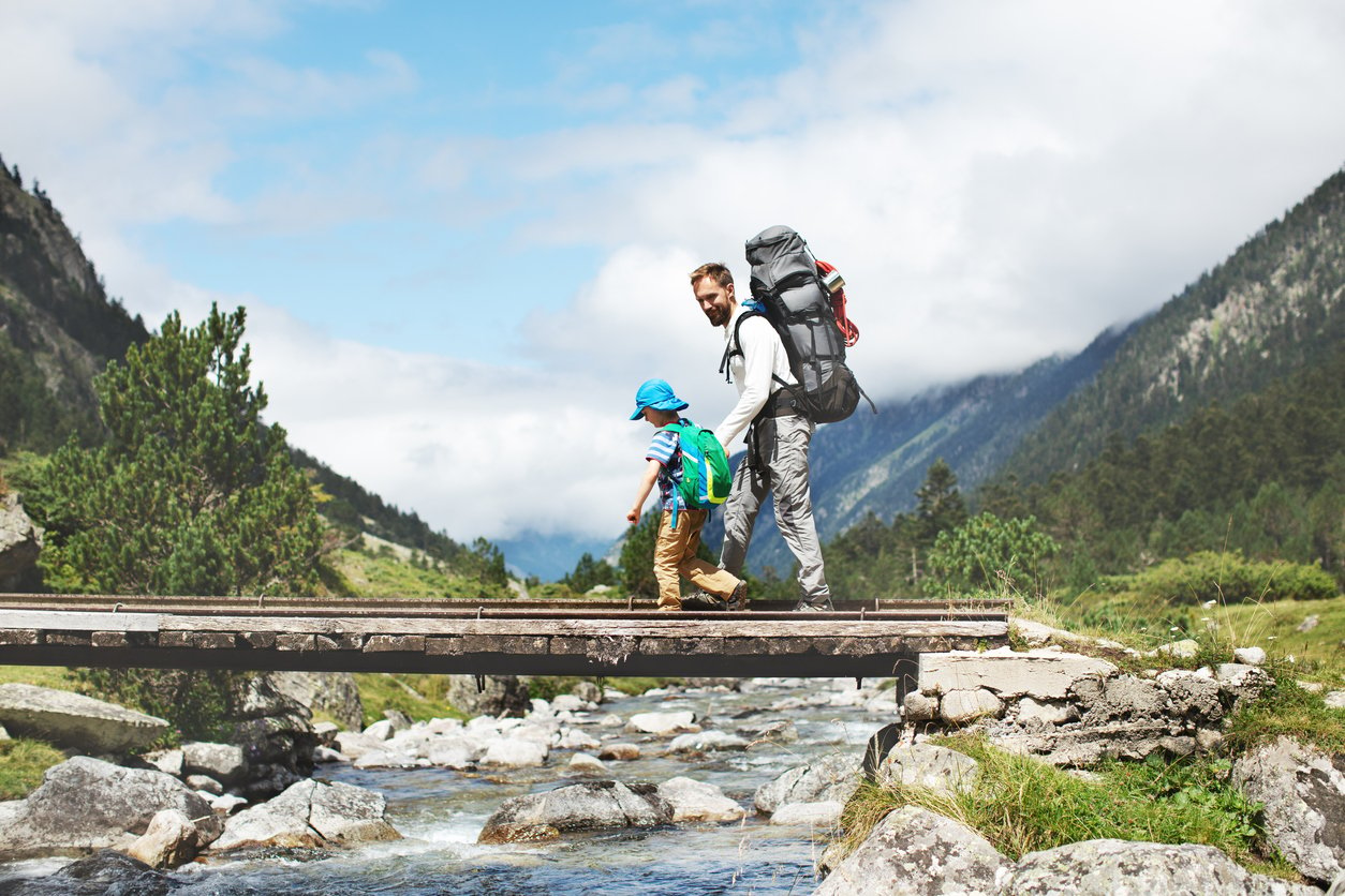 Father and son trekking in the mountains, crossing a bridge in front of lush green scenery.