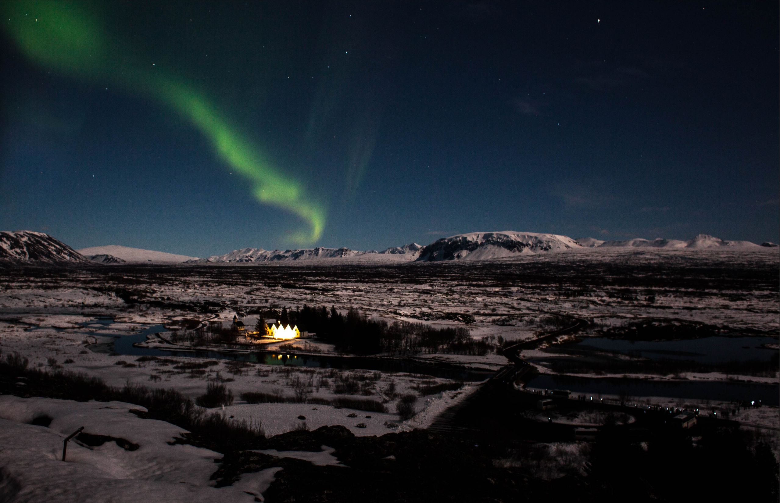 The northern lights blaze faintly above a snow covered landscape and a small village.