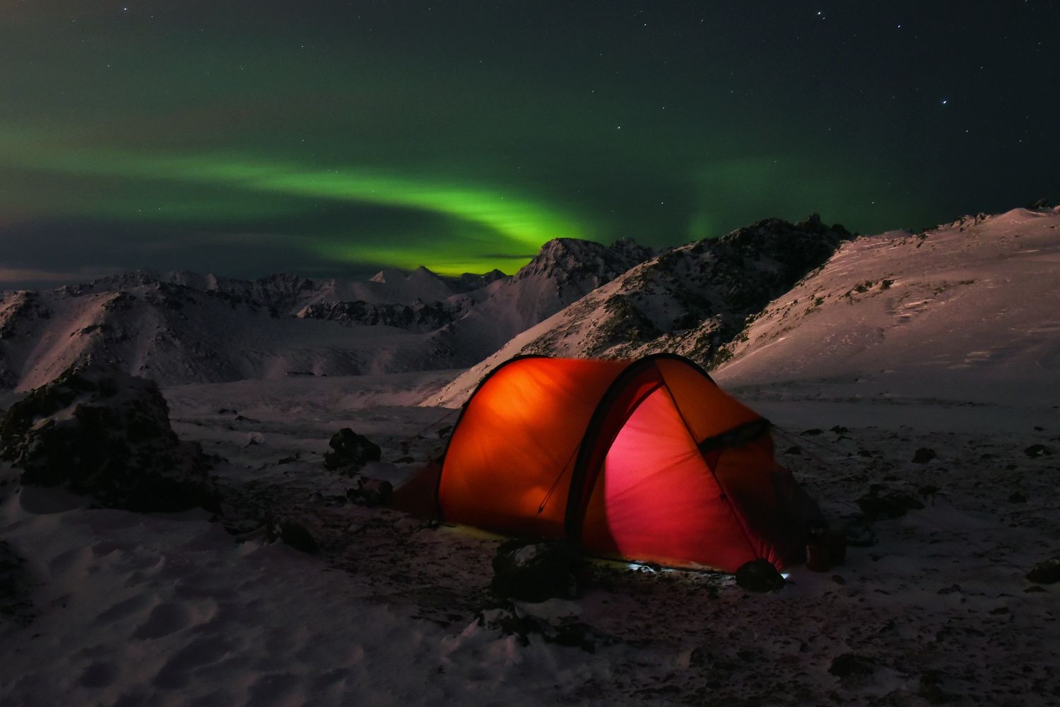 An illuminated tent in the snow, below the northern lights.