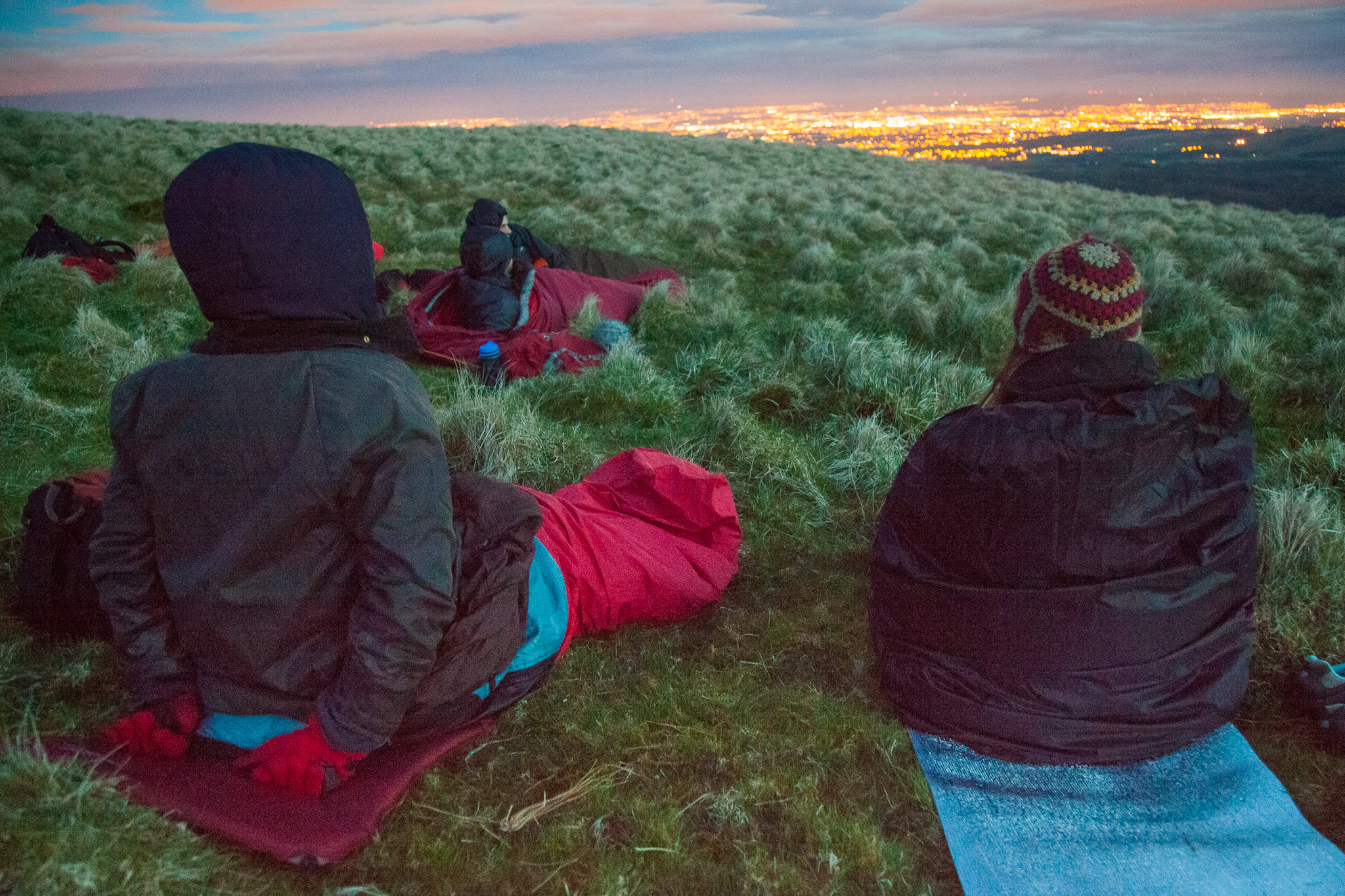 A group of people in sleeping bags on top of a hill, gazing at the illuminated city below.