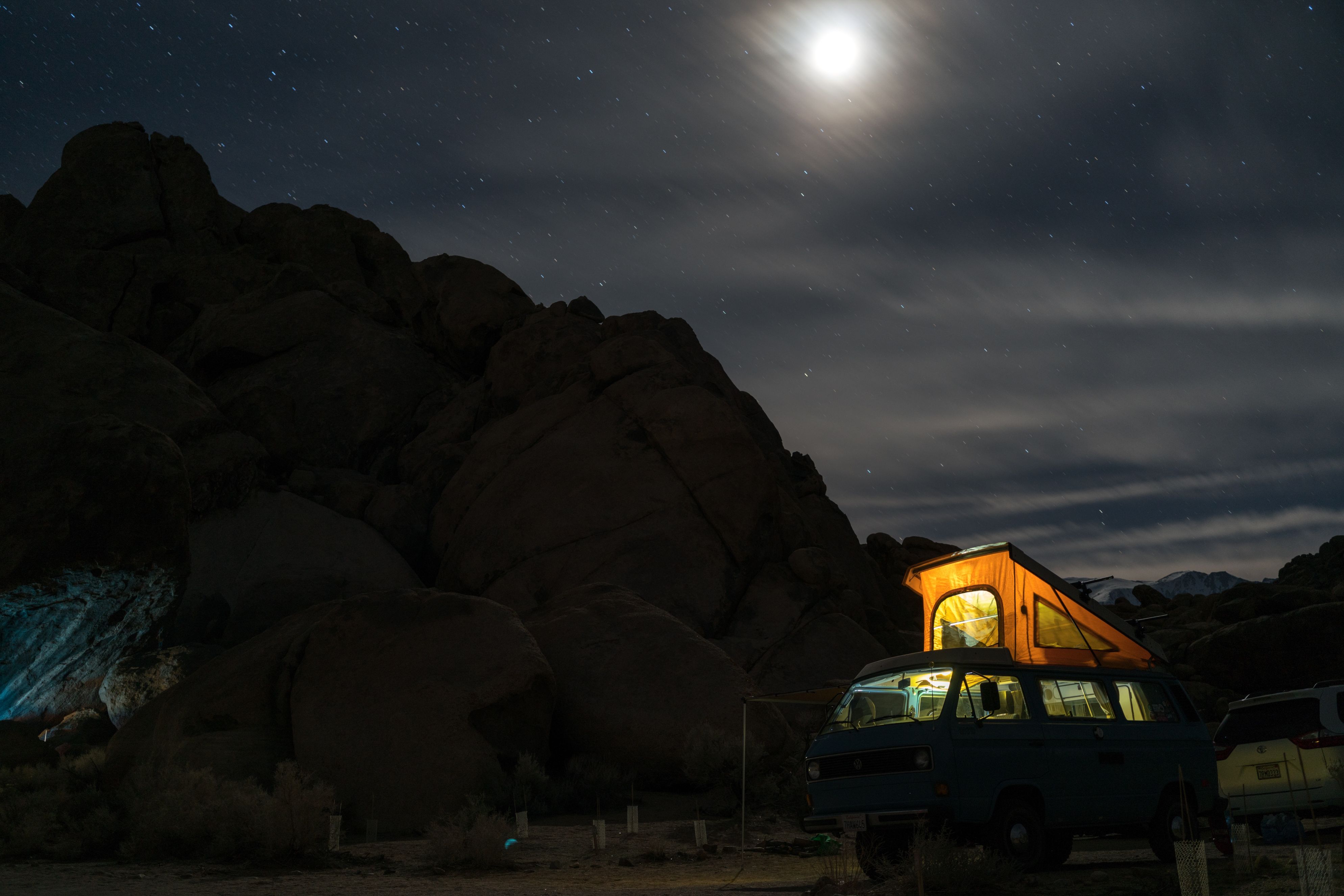 A camper van with a tent pitched on the roof sits below a mountain at night