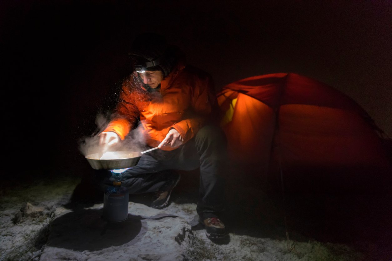 Someone cooking on a camping stove at night, by the light of a headtorch.
