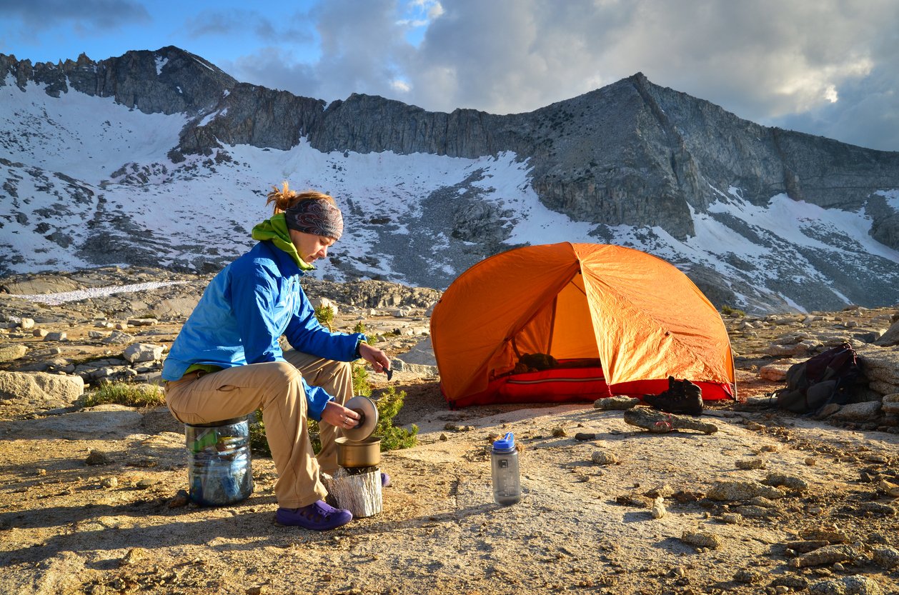 A woman cooking over a camping stove while wild camping, with tent and mountains in the background.