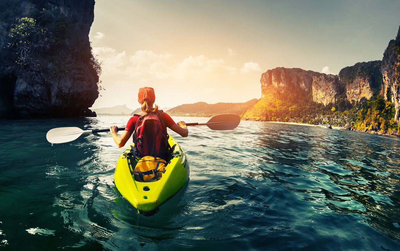 A woman kayaking, a rocky sunlit coastline in the background.