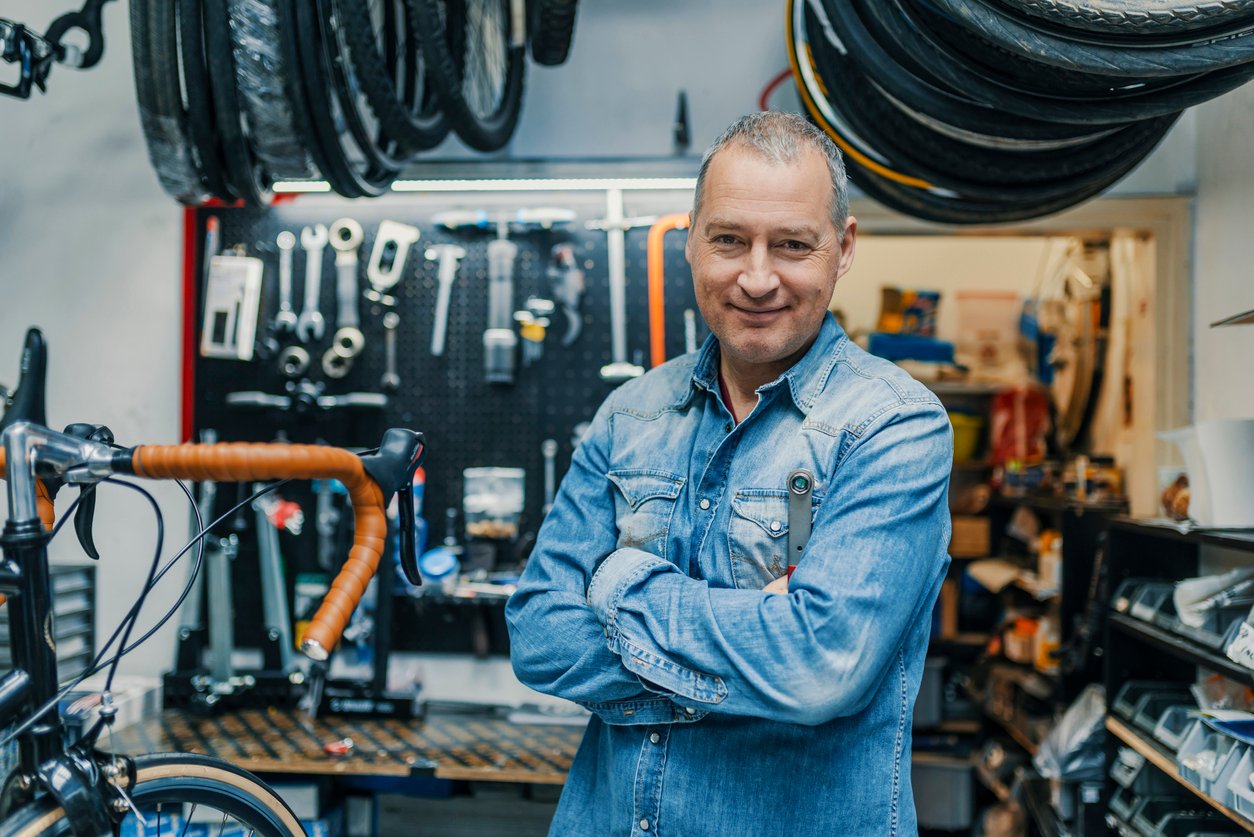 A man in a blue denim shirt stands in front of a bike workshop, smiling.