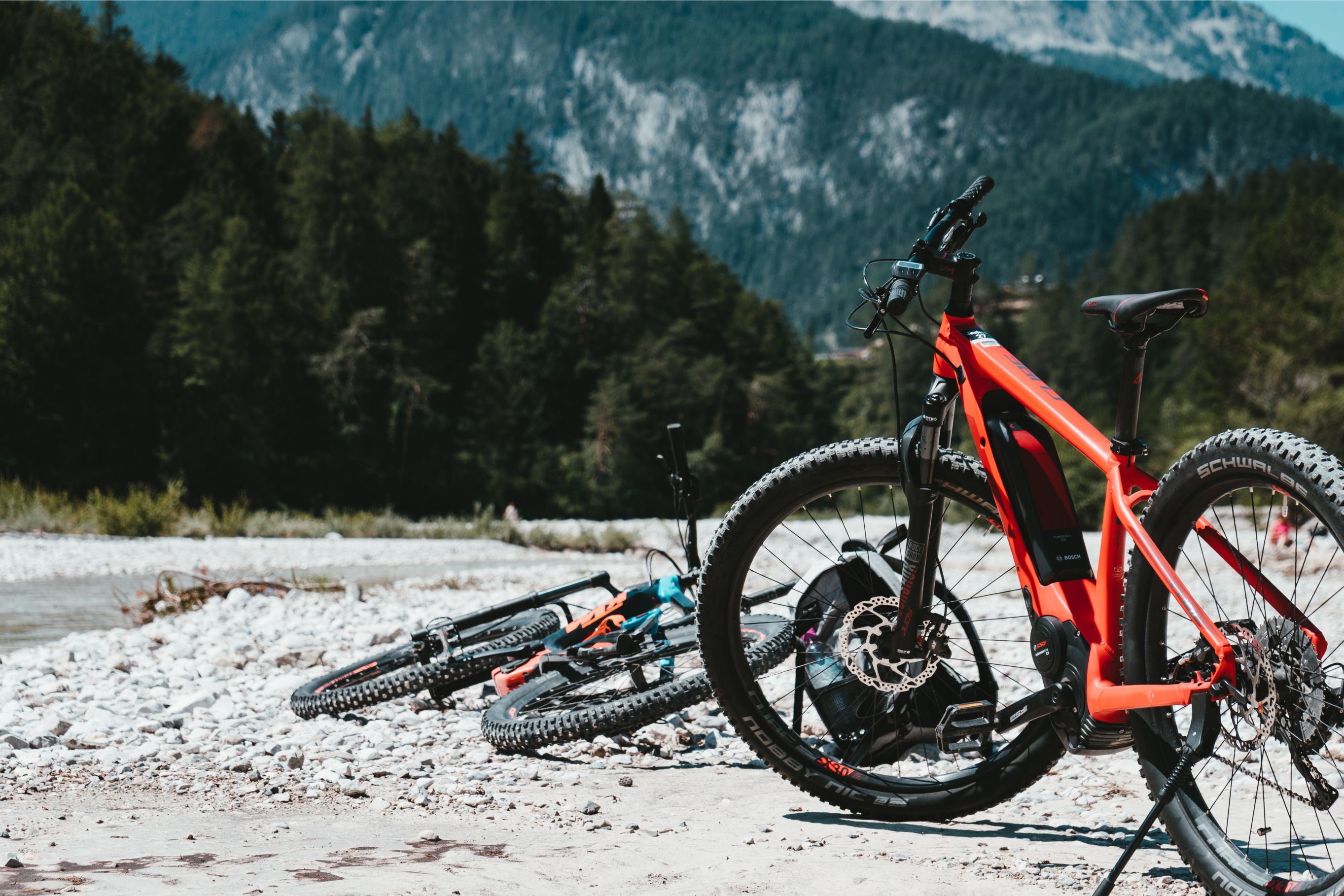 Three e-bikes sit by a riverside, with forests and mountains layering into the distance behind.