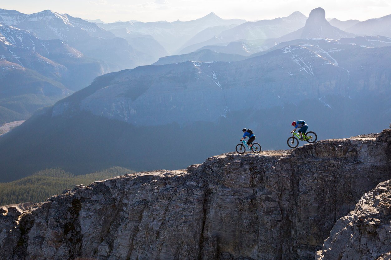 Two mountain bikers on the trail, a backdrop of mountains behind them.