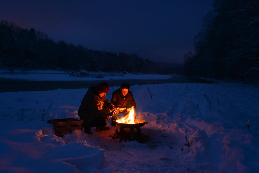 Two people huddled around a campfire in the snow.