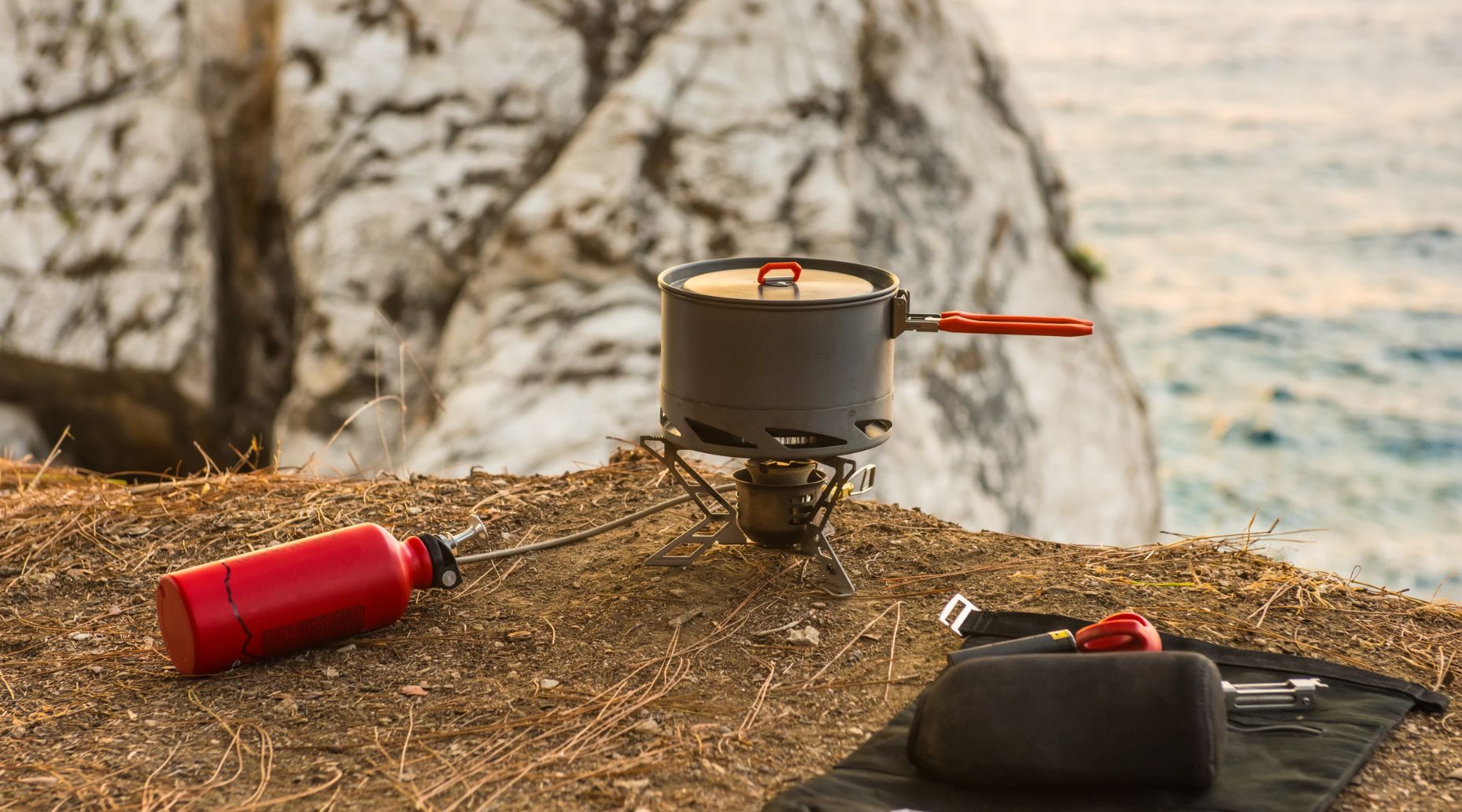 A camping stove with a saucepan on top, with cliffs and ocean in the background.