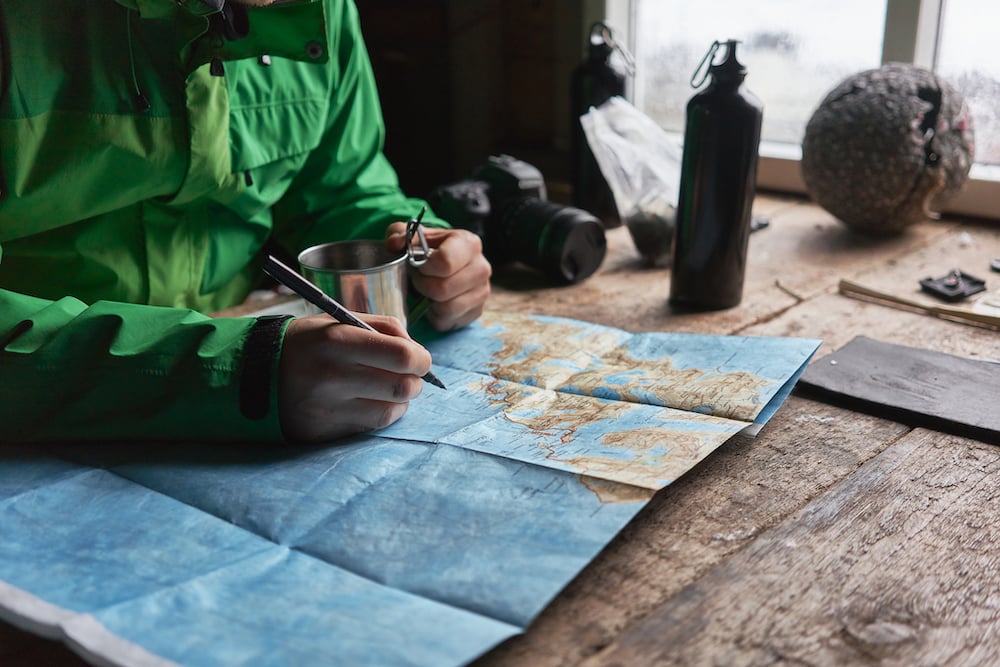 A hiker in a cabin makes notes on a map, while drinking coffee.