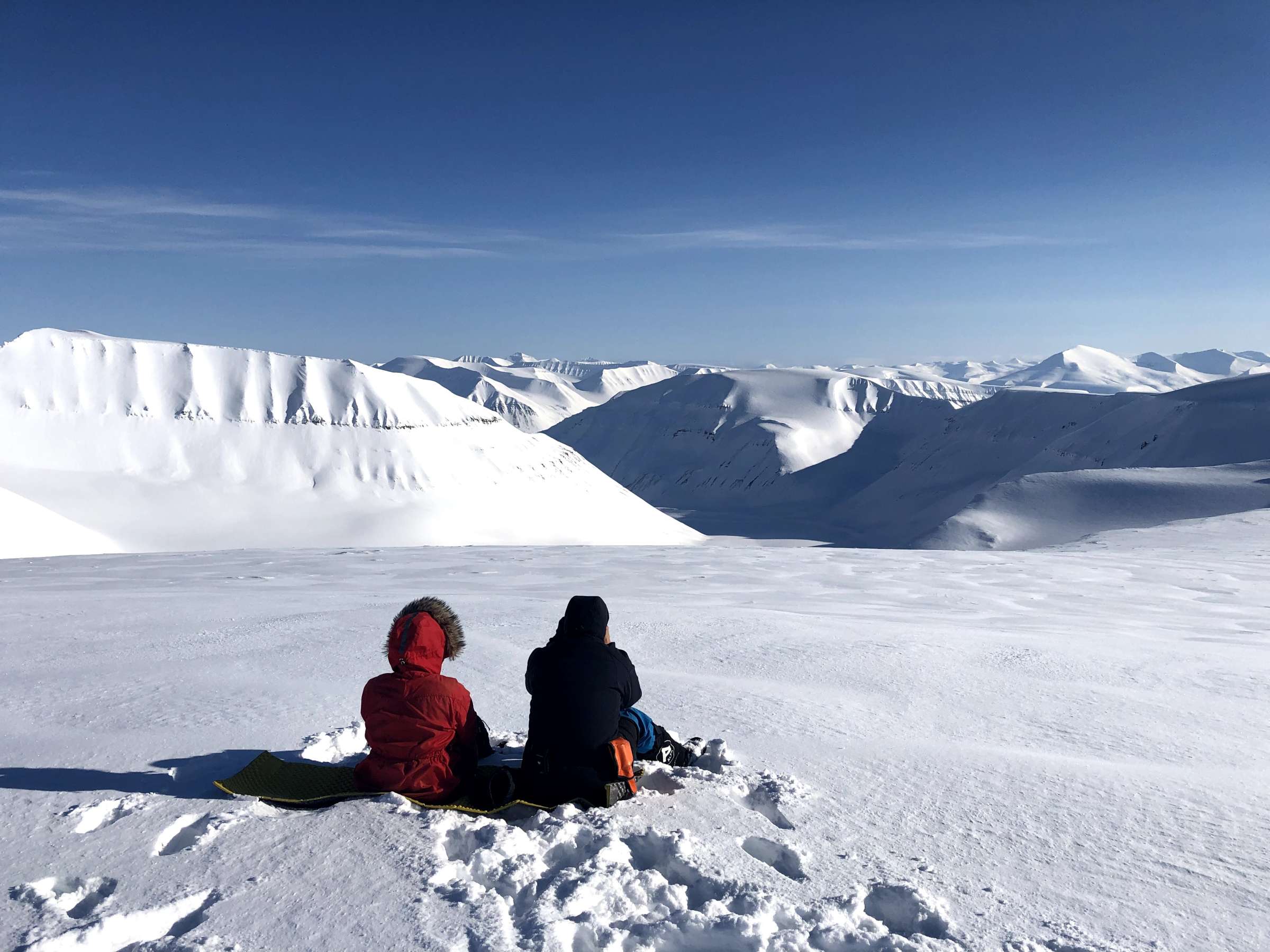 Two adventurers it on the snow of Svalbard, looking out over the environment.