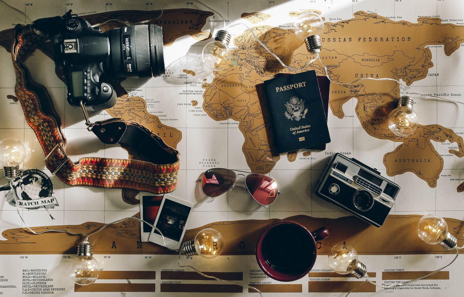 A scratch map in the background; a camera, passport and other travel accessories are on top of it.