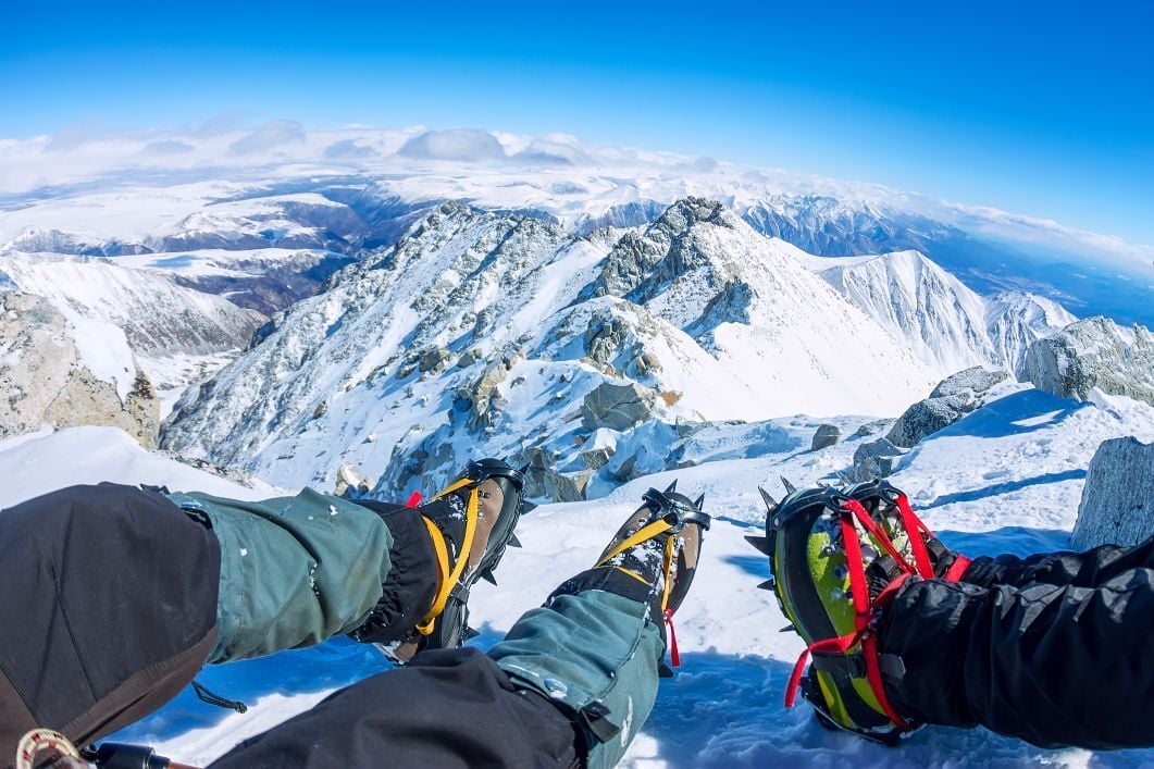 Two hikers show off their crampons on the summit of an icy mountain.