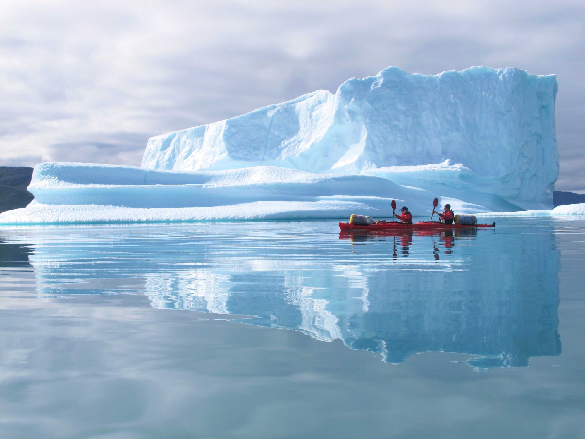 Two kayakers in a double kayak in the shadow of an enormous iceberg in Greenland.