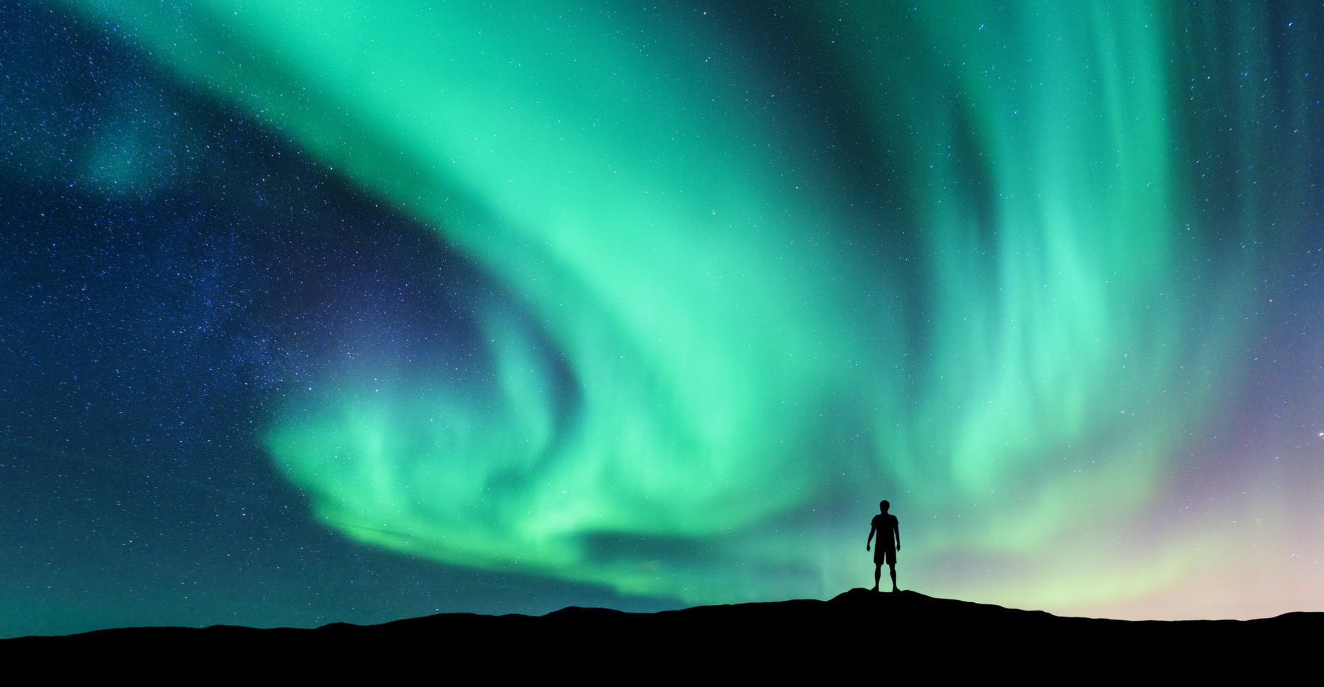 A small figure silhouetted in front of a spectacular green aurora borealis display.