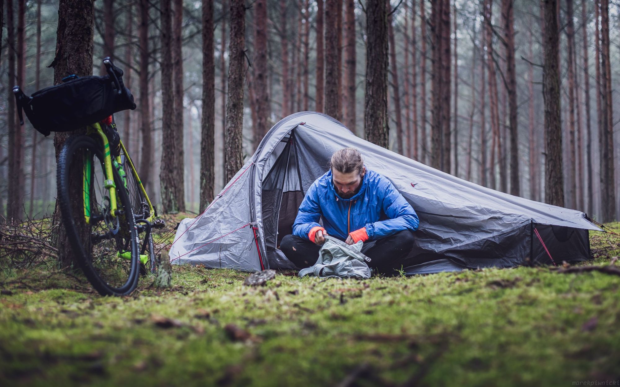 A camper sitting outside his tent in a forest, with a bike on the left hand side.
