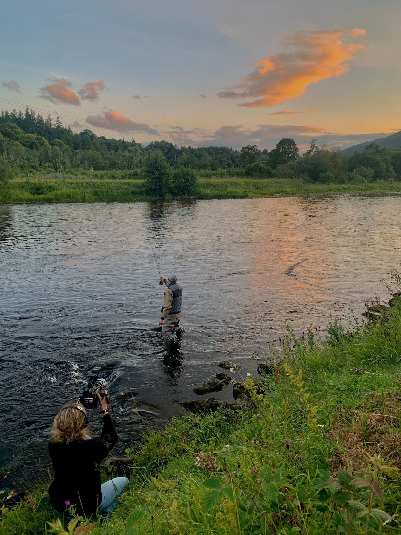 A woman filming a fisherman standing in the stream in the Scottish Highlands.