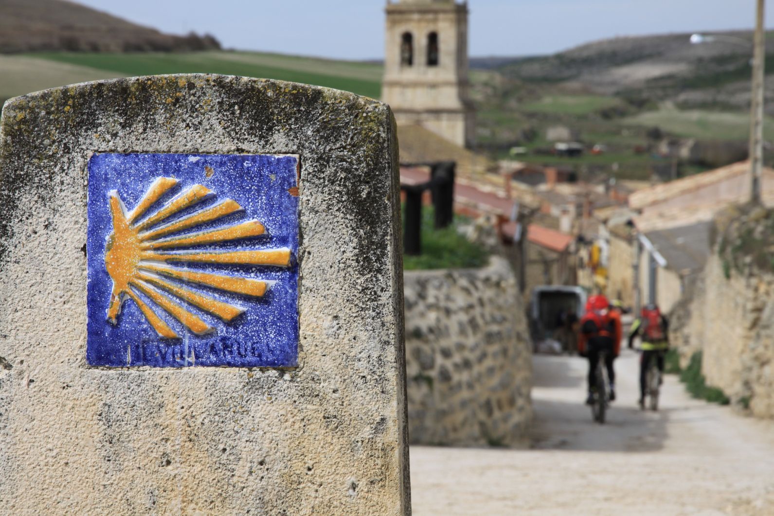 A Camino waymark in the foreground; hikers and a hill village in the background.