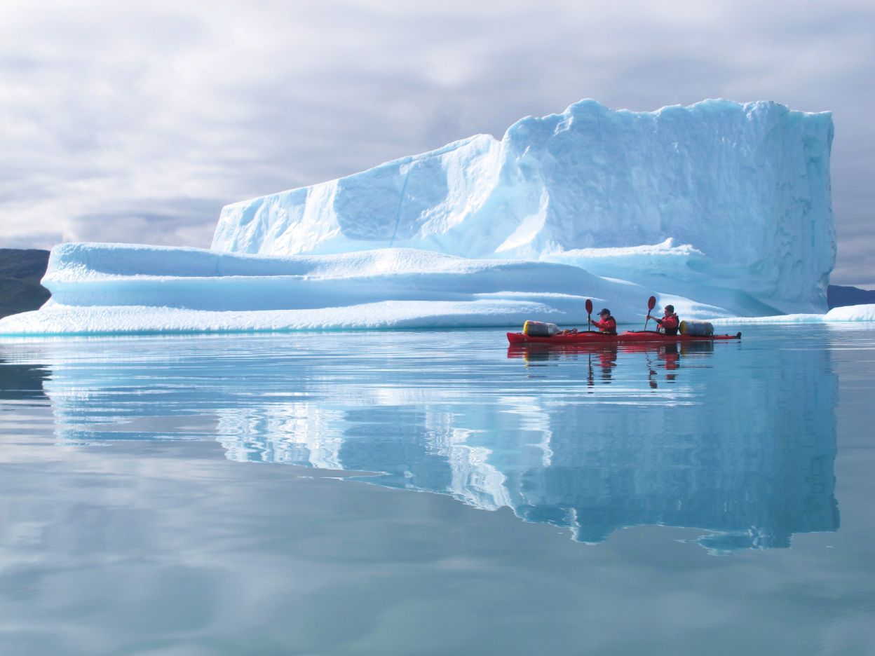 Two people kayak past a large iceberg in Greenland.