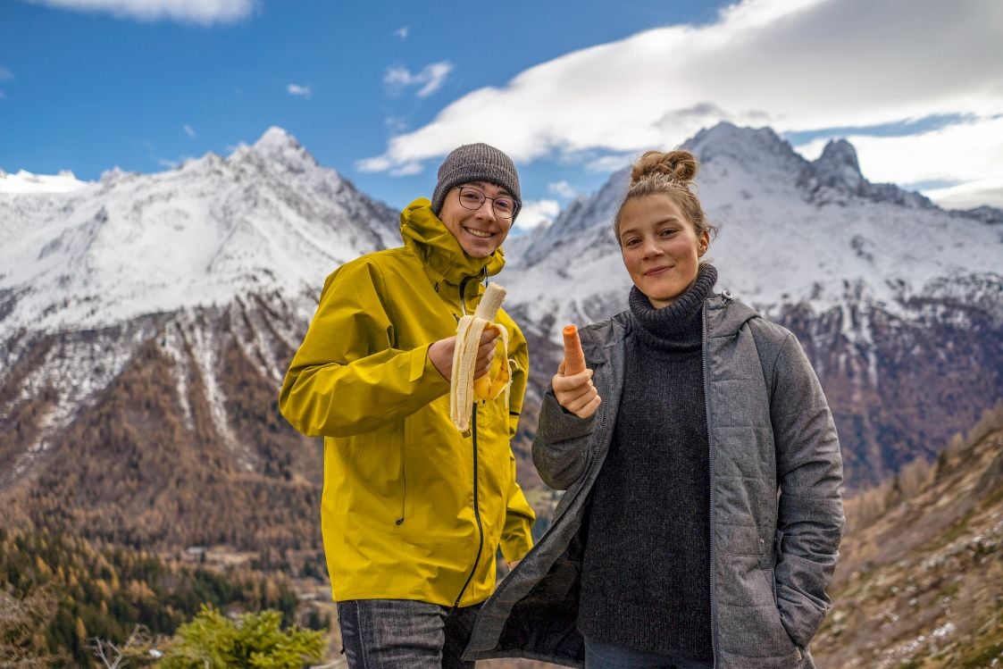 Two hikers; one eating a banana and the other eating a carrot.