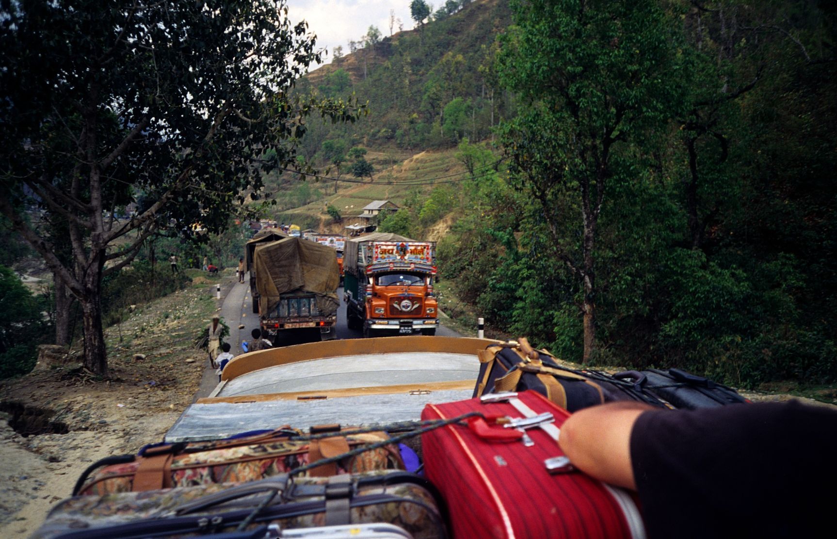 Close up of someone sitting on top of a bus filled with luggage, in Nepal - lorries on the road in the background.
