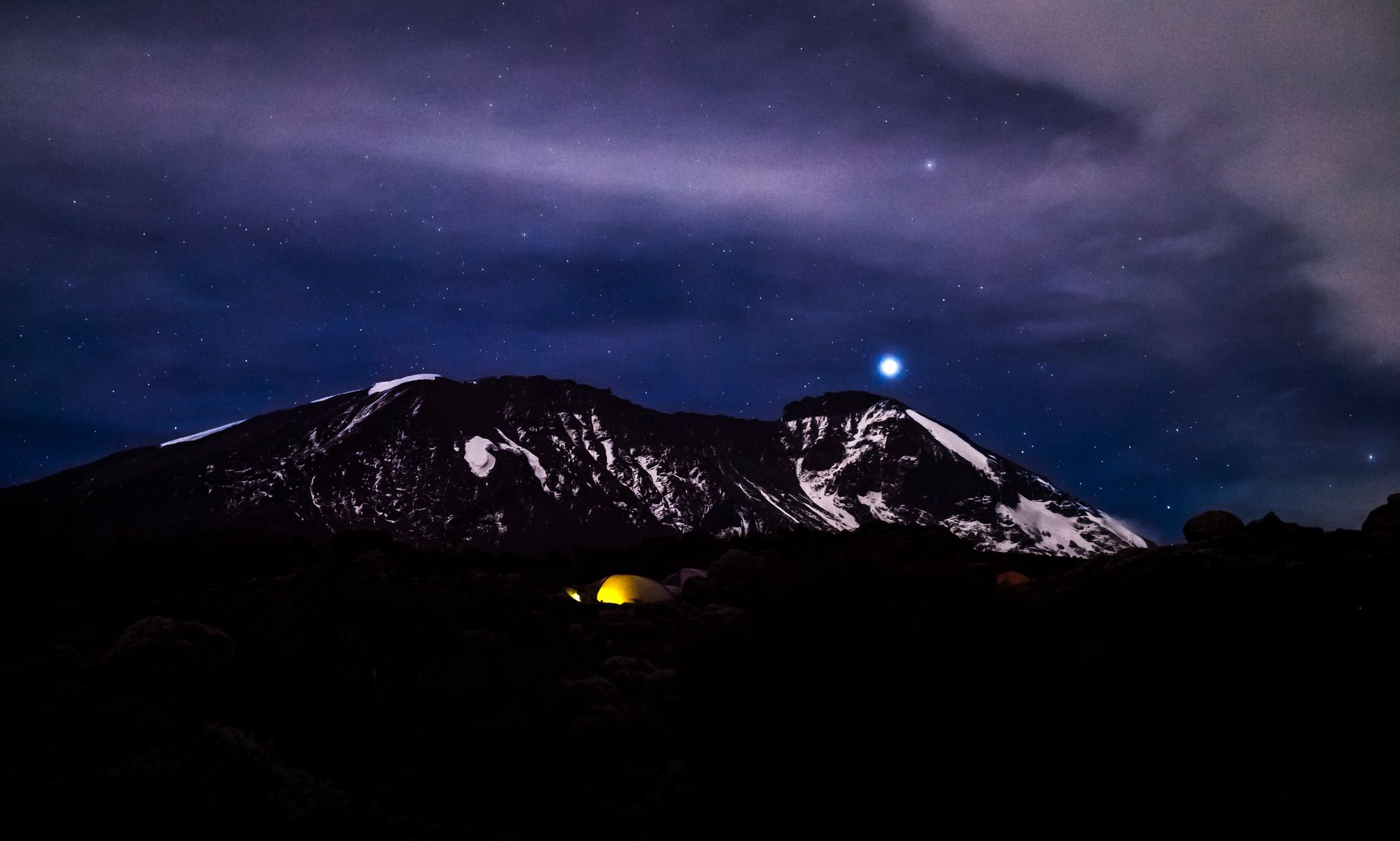 An illuminated tent camped on Kilimanjaro - it's nightfall and there's a cold blue moon overhead.